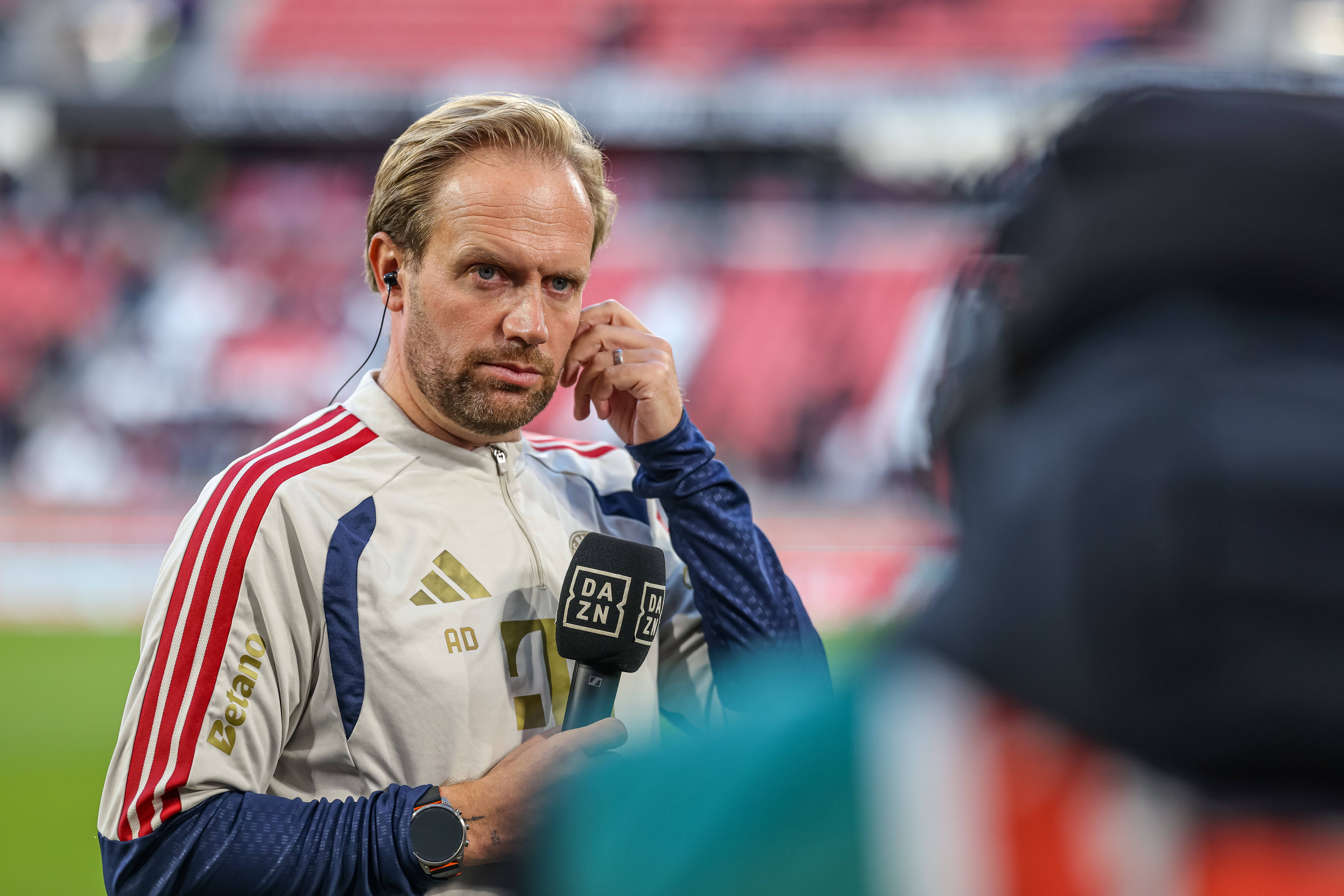 Ein Mann im Trainingsanzug spricht mit einem DAZN-Mikrofon auf einem Fußballfeld.