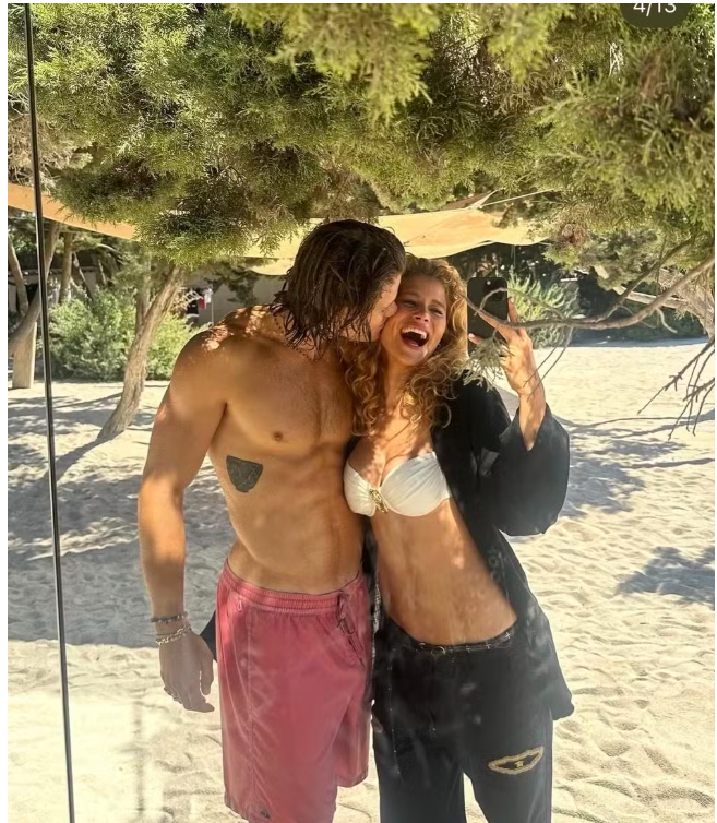 Ein Paar posiert lachend am Sandstrand unter Bäumen, sie machen ein Selfie.