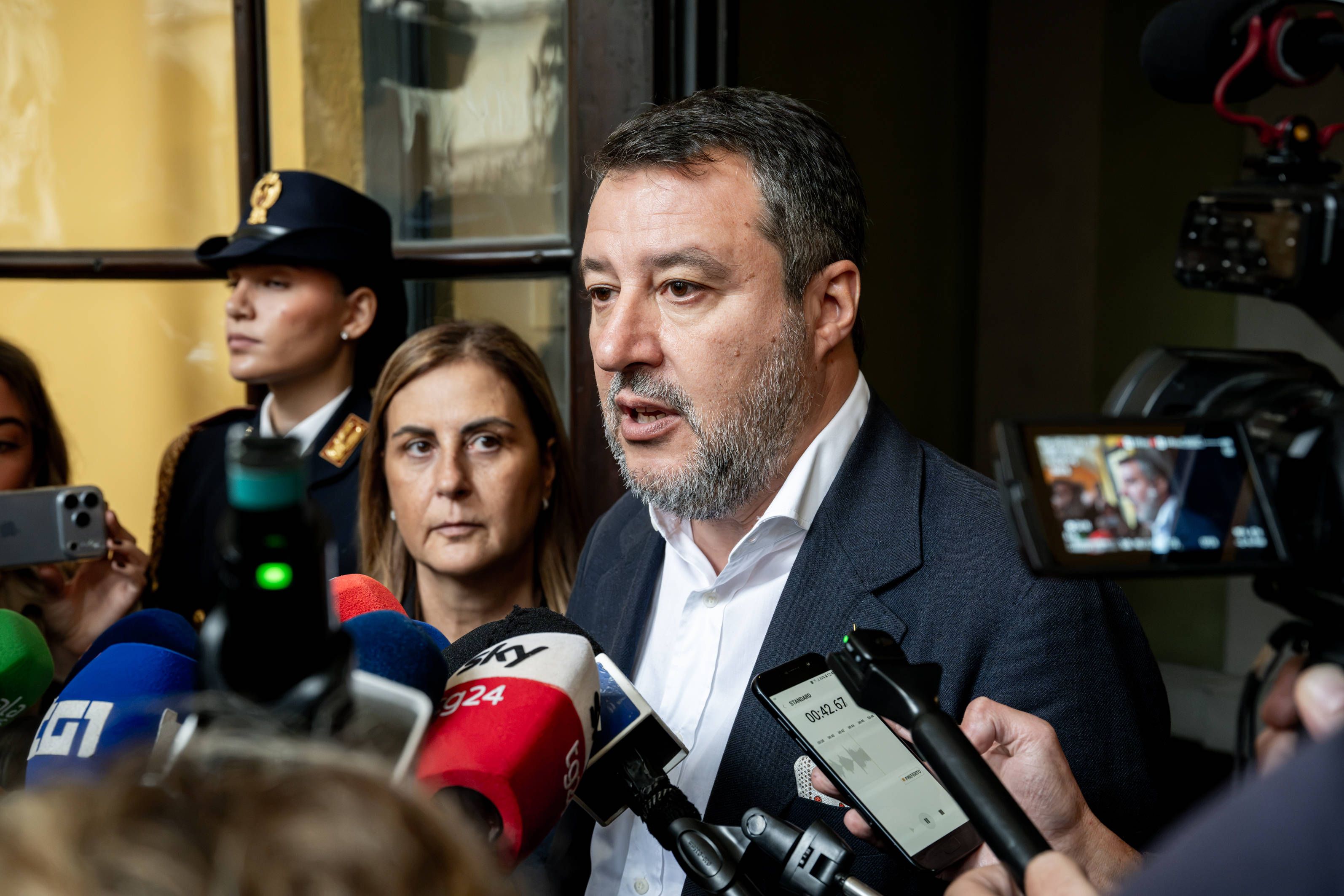 Ein Mann spricht vor Mikrofonen und Kameras bei einer Pressekonferenz, mehrere Menschen stehen um ihn.