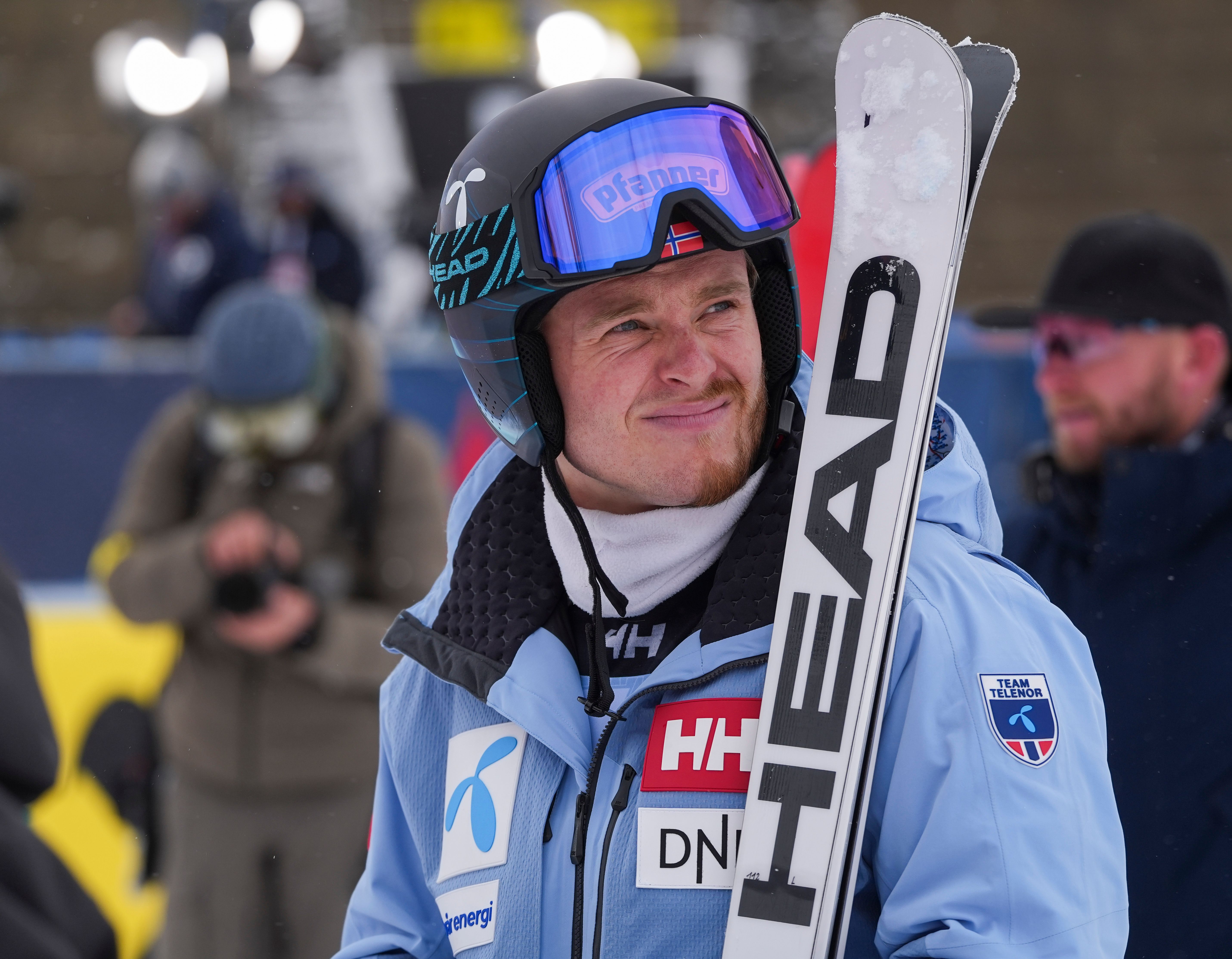 Ein Skifahrer im blauen Anzug hält Ski und trägt Helm mit Skibrille bei einem Wettkampf.