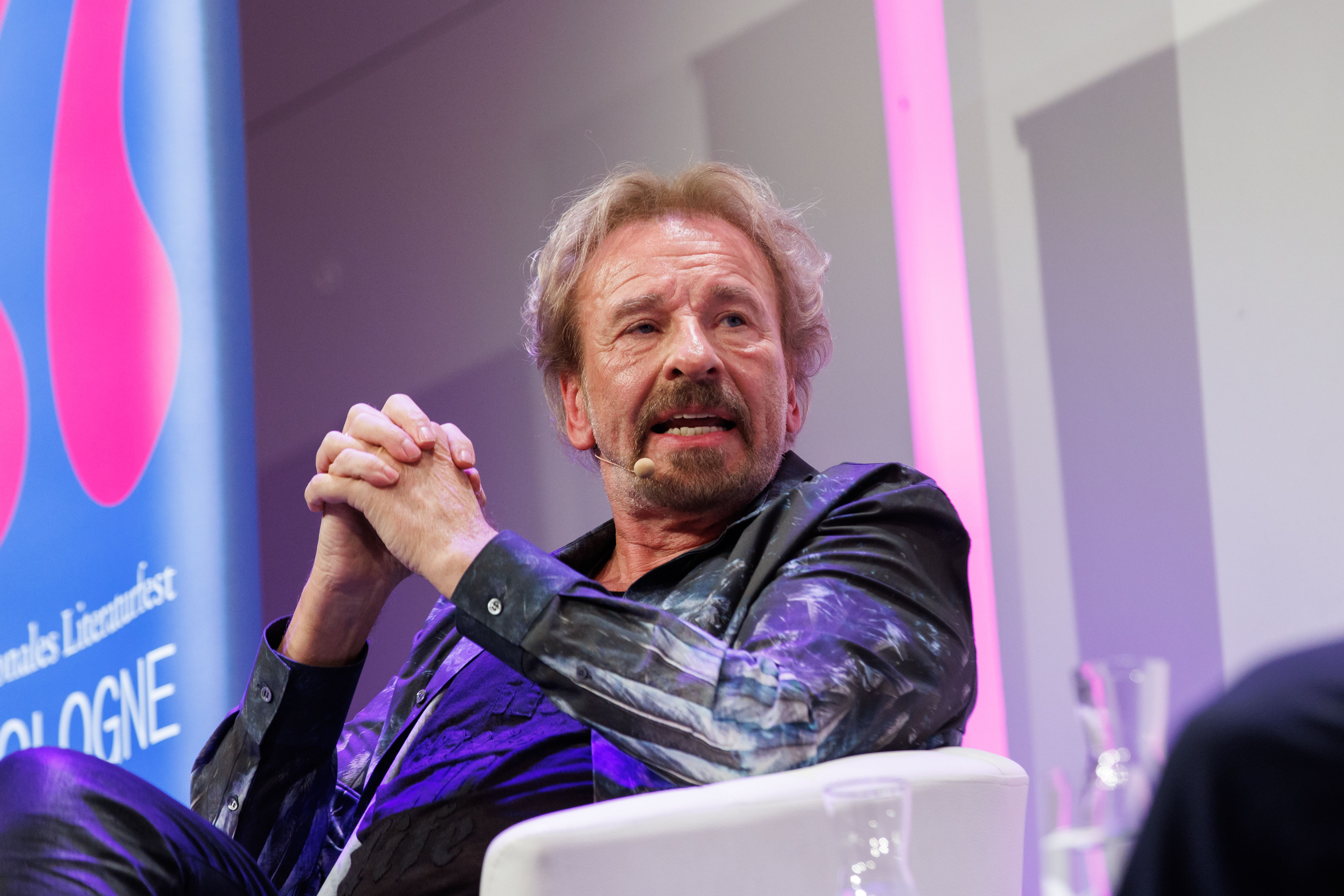 Ein Mann mit Mikrofon spricht auf einer Bühne, im Hintergrund bunte Beleuchtung und Banner.