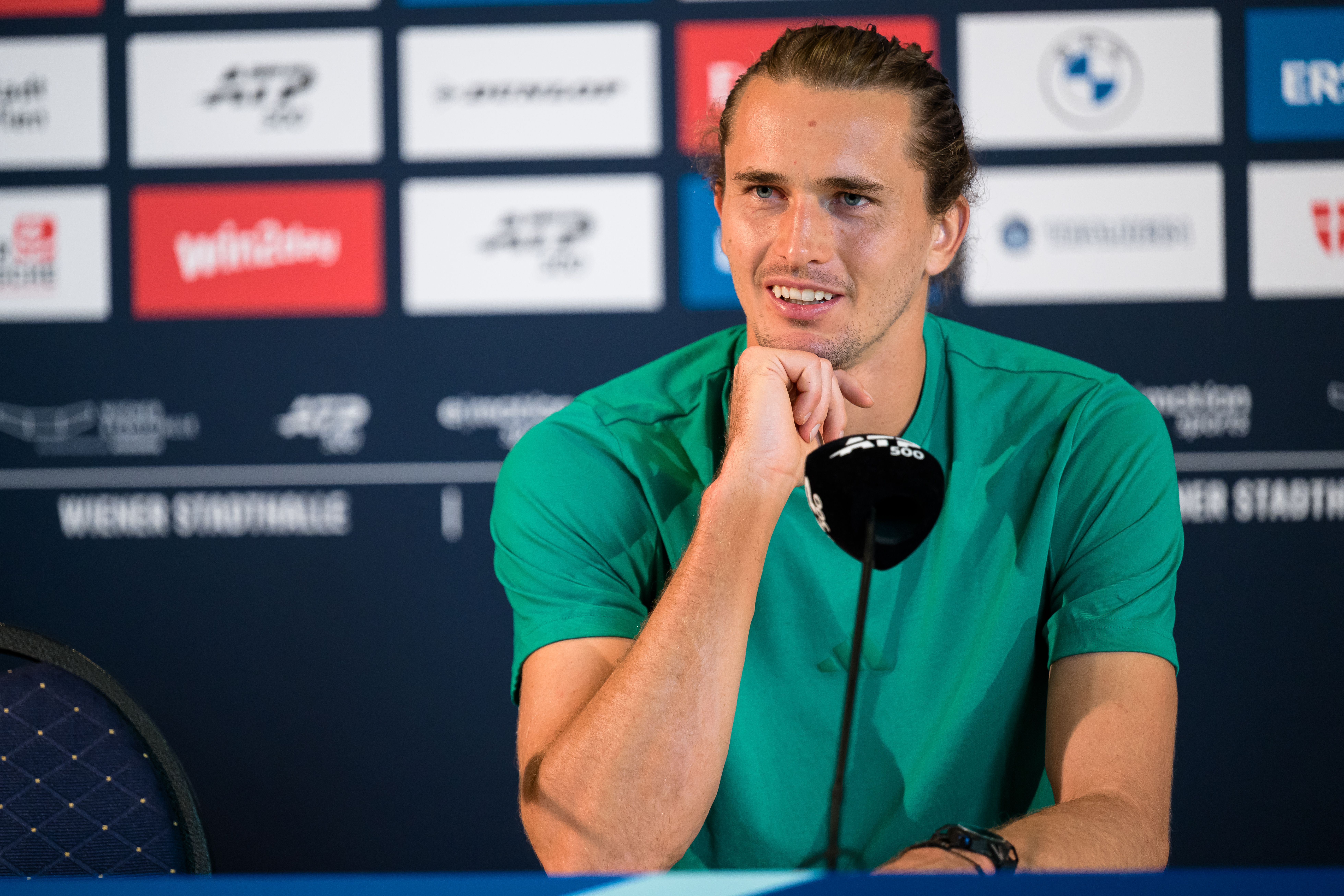 Ein Mann im grünen Shirt spricht lächelnd bei einer Pressekonferenz vor einem Mikrofon.
