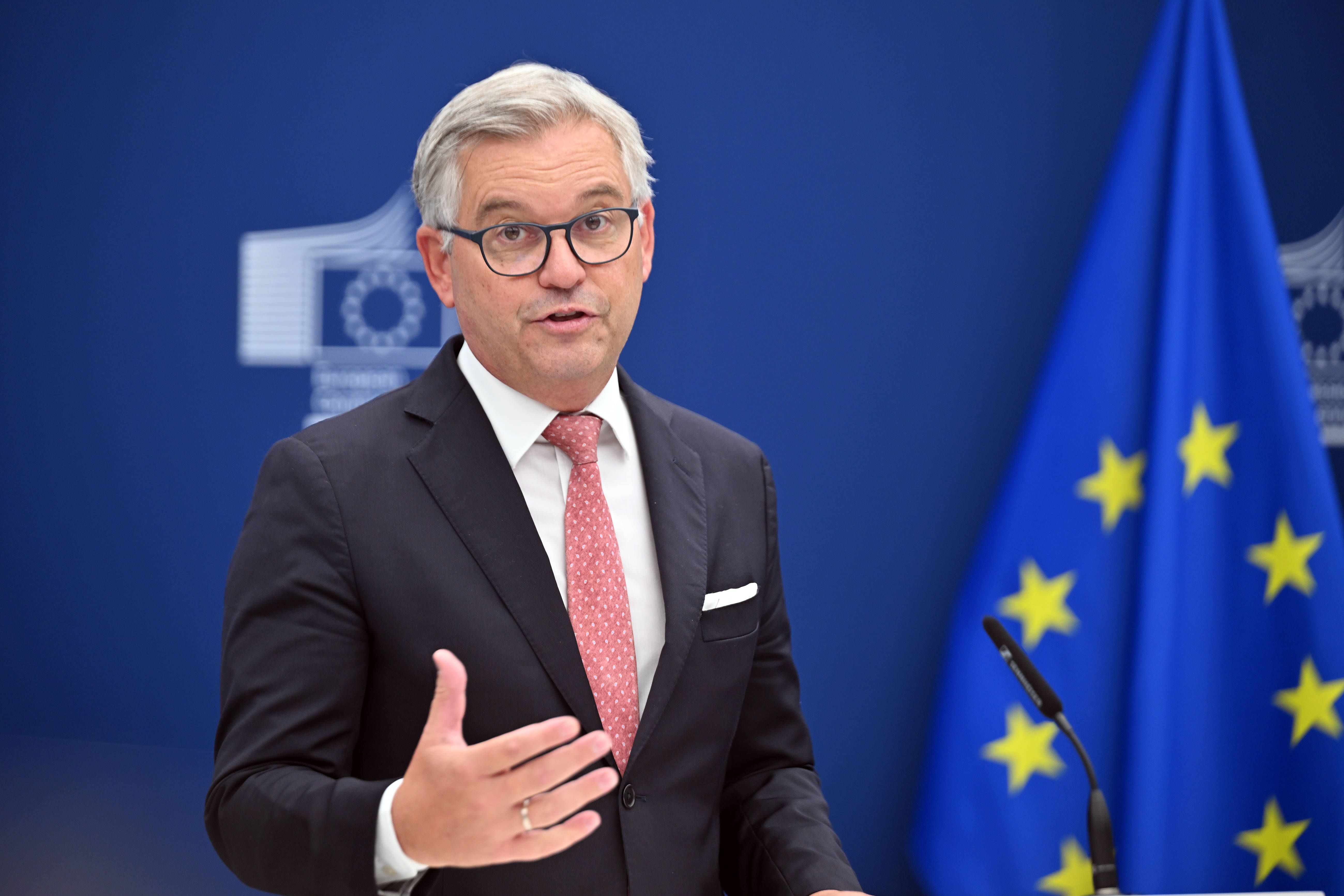 Ein Mann im Anzug spricht auf einer Pressekonferenz vor einer EU-Flagge und blauem Hintergrund.