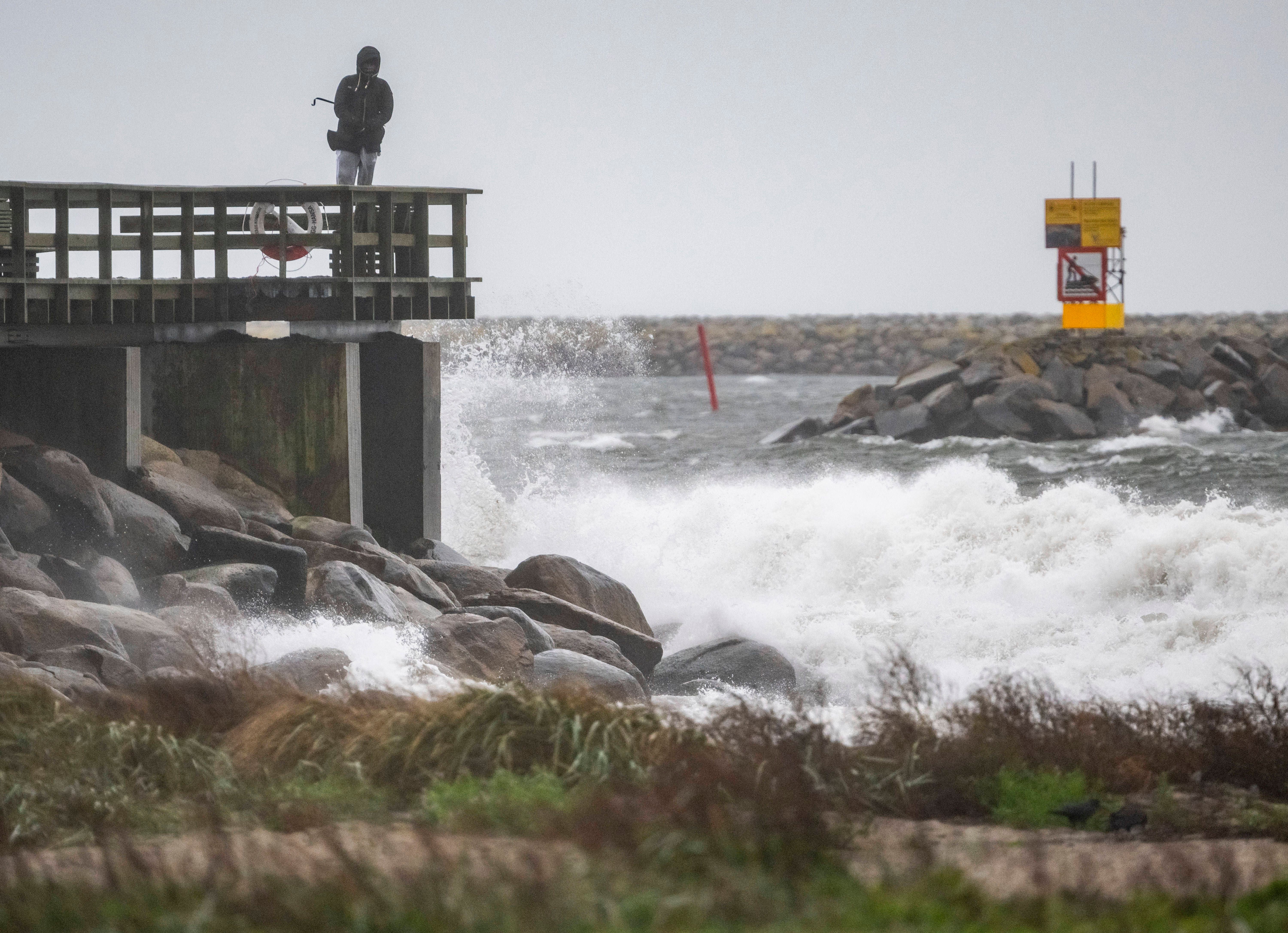 Eine Person steht auf einem Pier und beobachtet hohe Wellen bei Sturmwind an der Küste.
