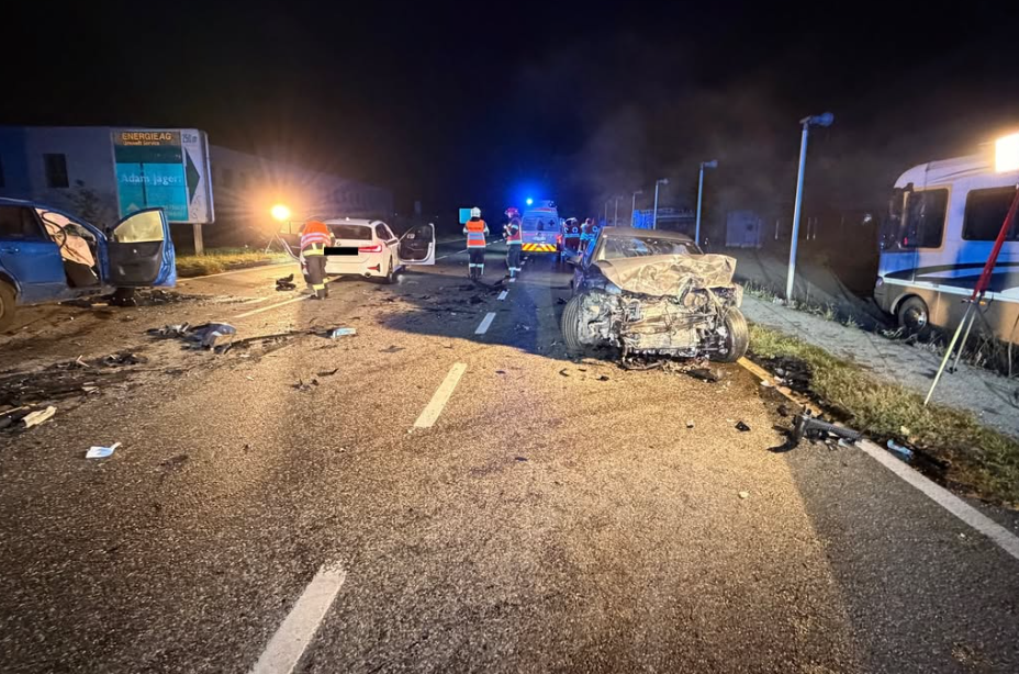 Nachtszene eines schweren Autounfalls mit mehreren beschädigten Fahrzeugen und Einsatzkräften auf der Straße.