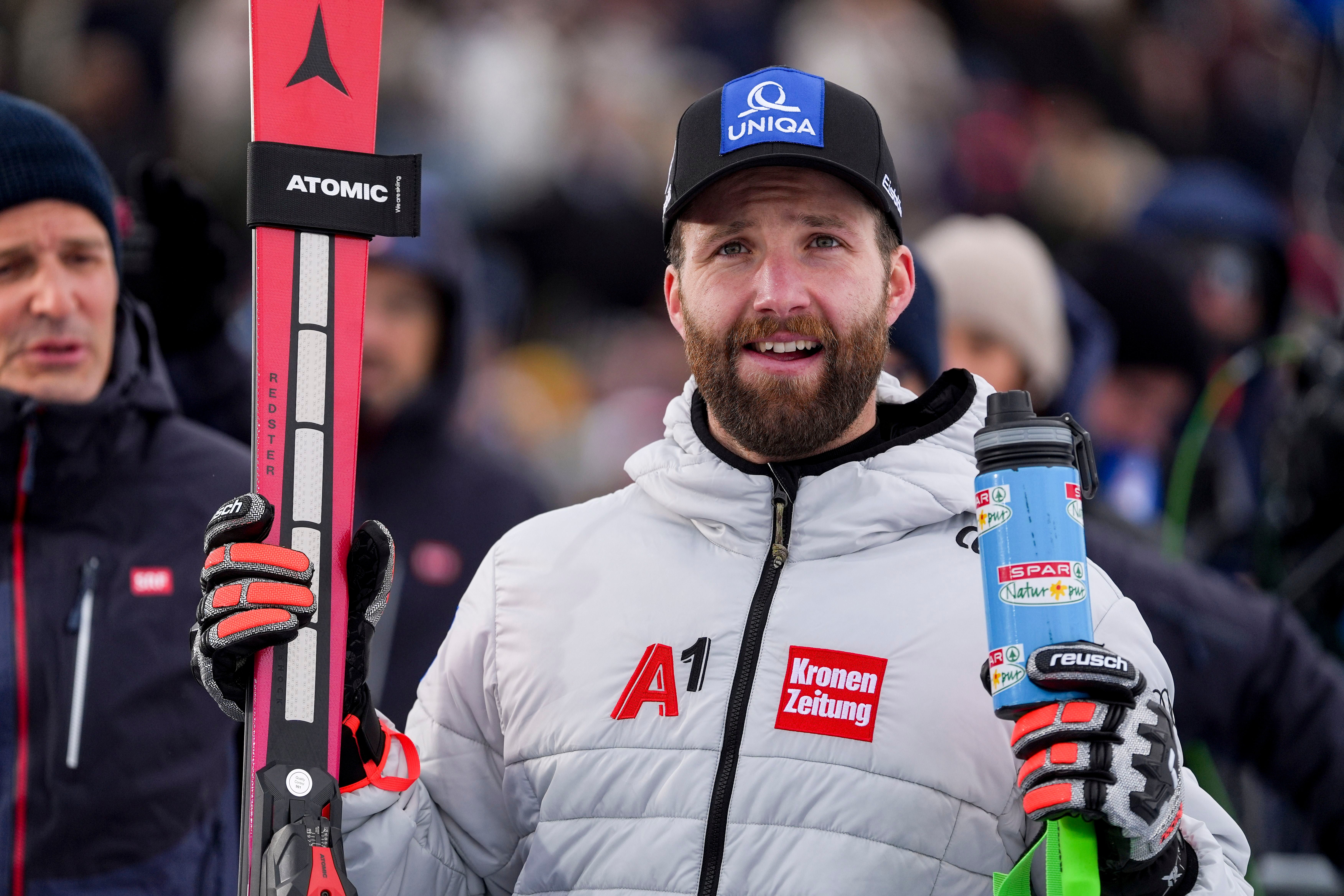 Ein Skifahrer im weißen Anorak hält Ski und Trinkflasche, Menschenmenge im Hintergrund.