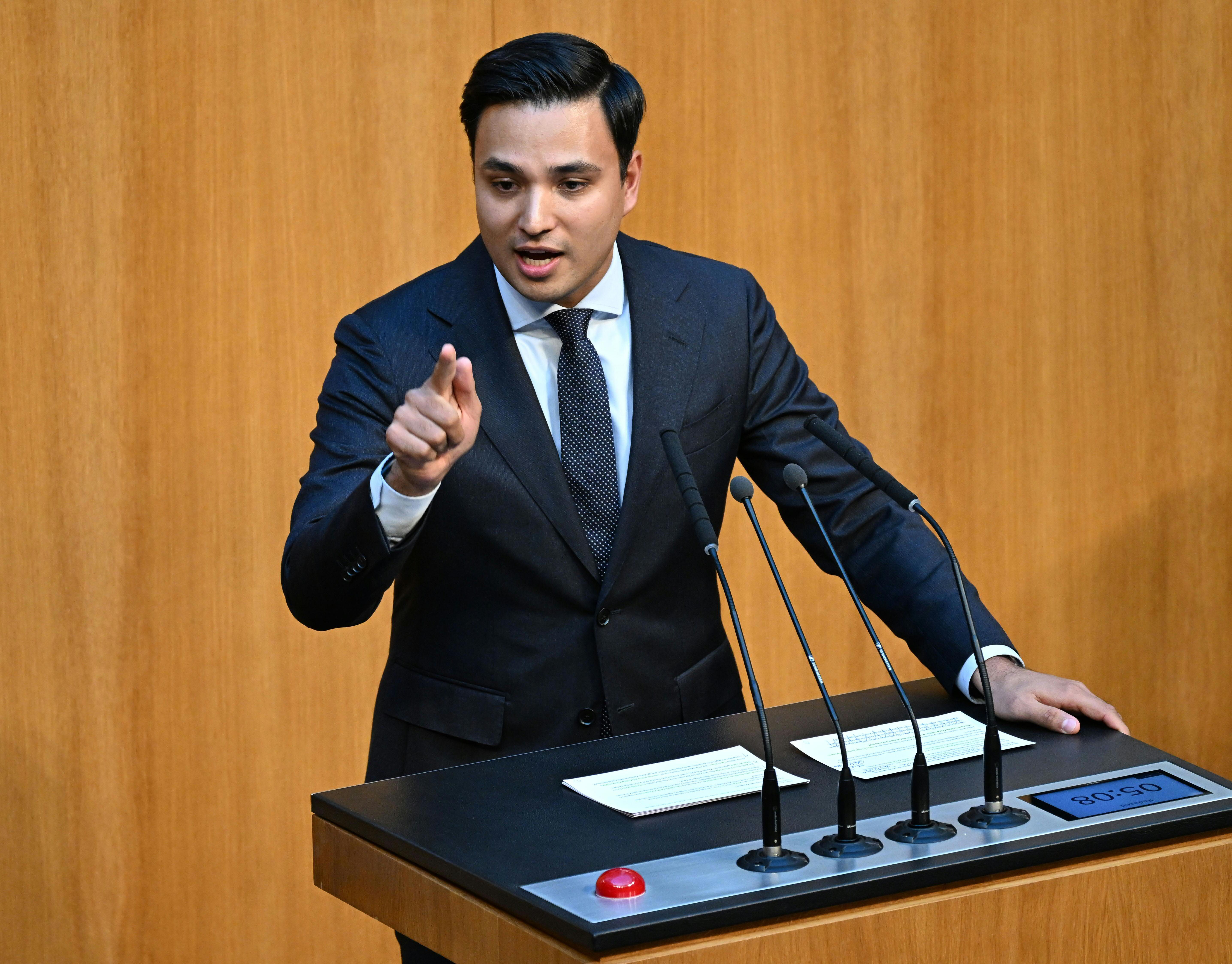 Ein Mann im Anzug spricht gestikulierend an einem Rednerpult mit Mikrofonen im Parlament.