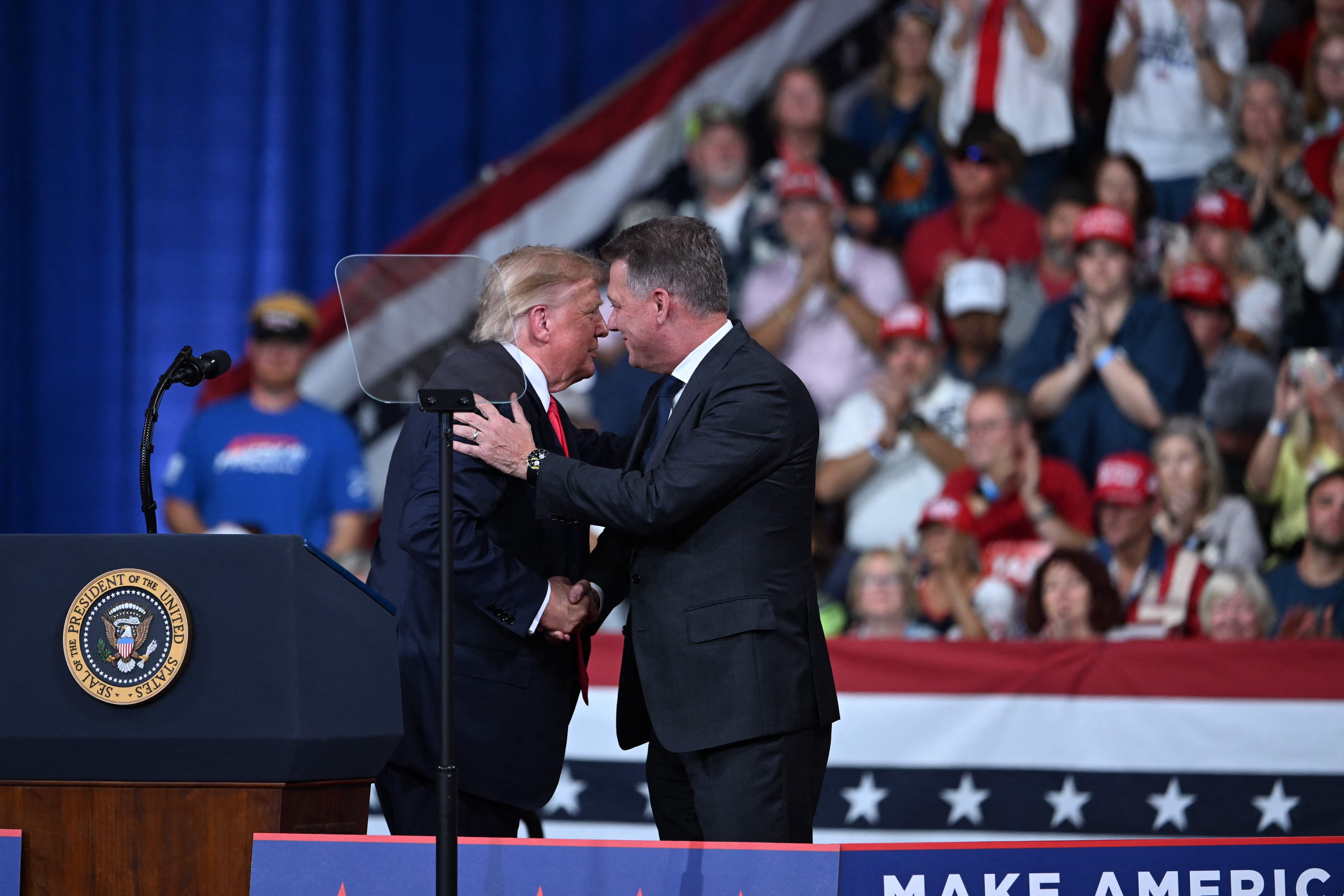 Donald Trump begrüßt einen Mann auf einer Wahlkampfveranstaltung mit jubelndem Publikum im Hintergrund.