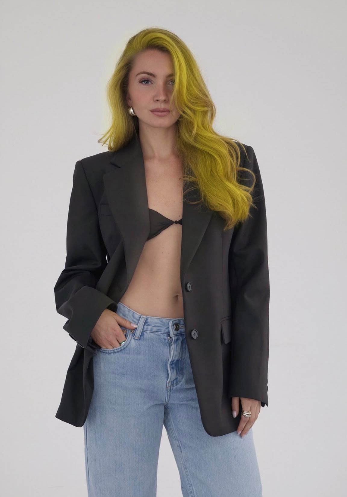 Frau mit gelben Haaren, schwarzem Blazer, BH und Jeans vor hellem Hintergrund.