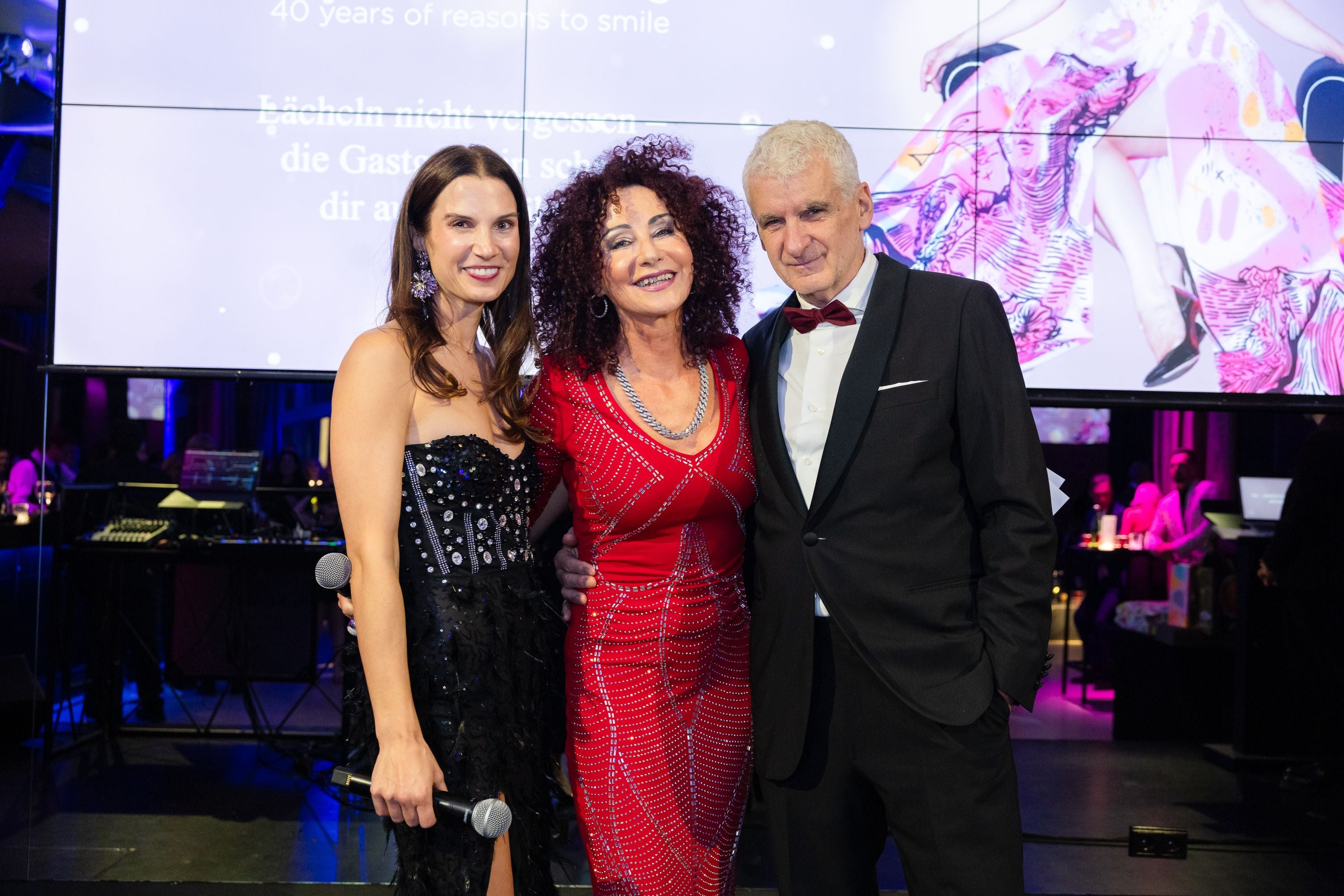 Drei elegant gekleidete Personen posieren lächelnd bei einer festlichen Veranstaltung.