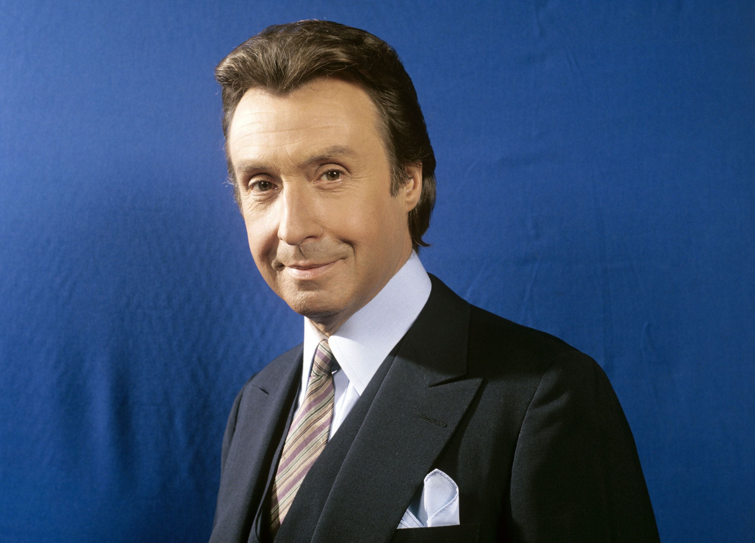 Ein Mann im Anzug und mit Krawatte steht vor blauem Hintergrund und lächelt in die Kamera.