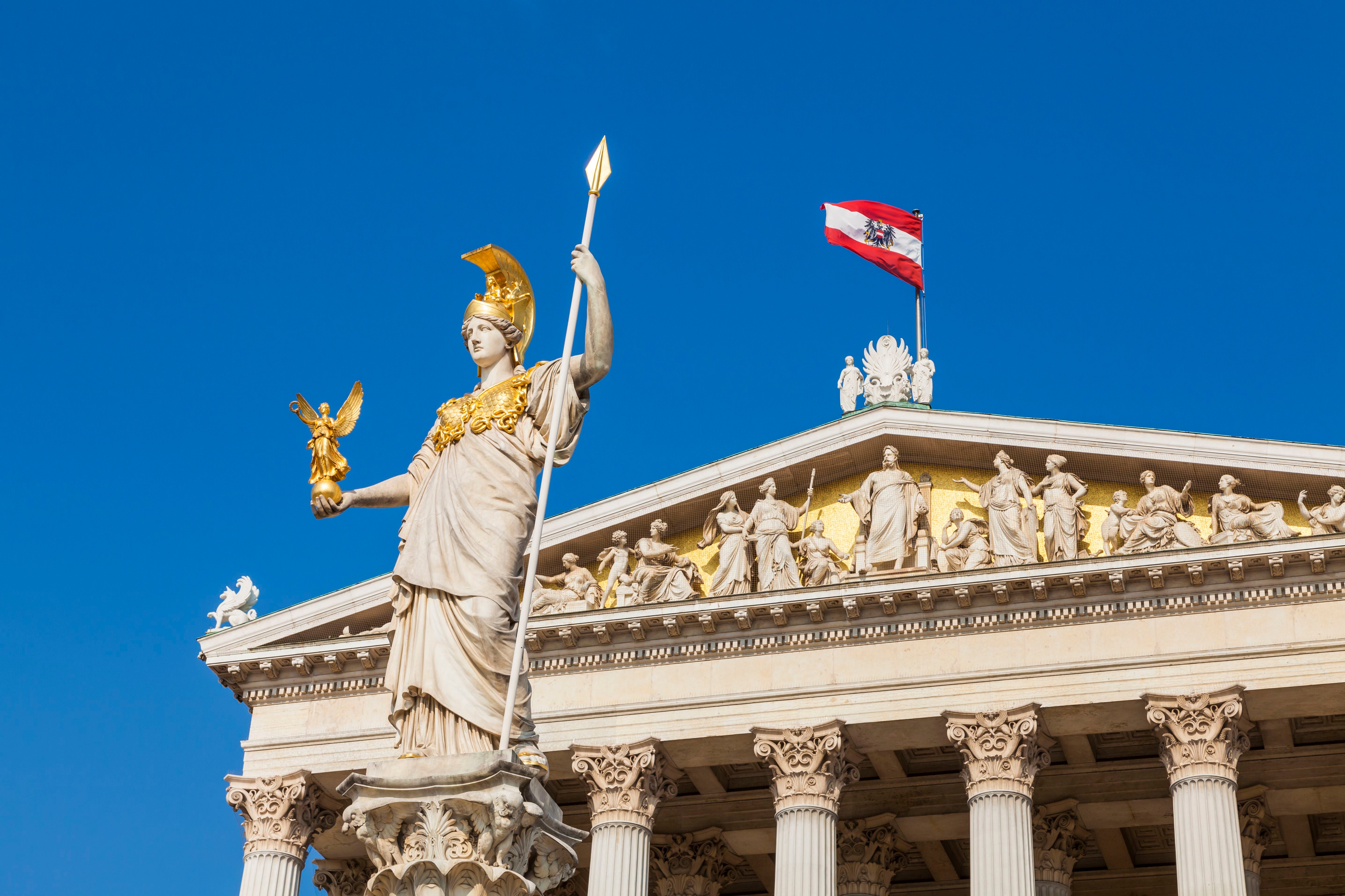 Statue vor dem österreichischen Parlament mit Nationalflagge auf dem Dach bei blauem Himmel.