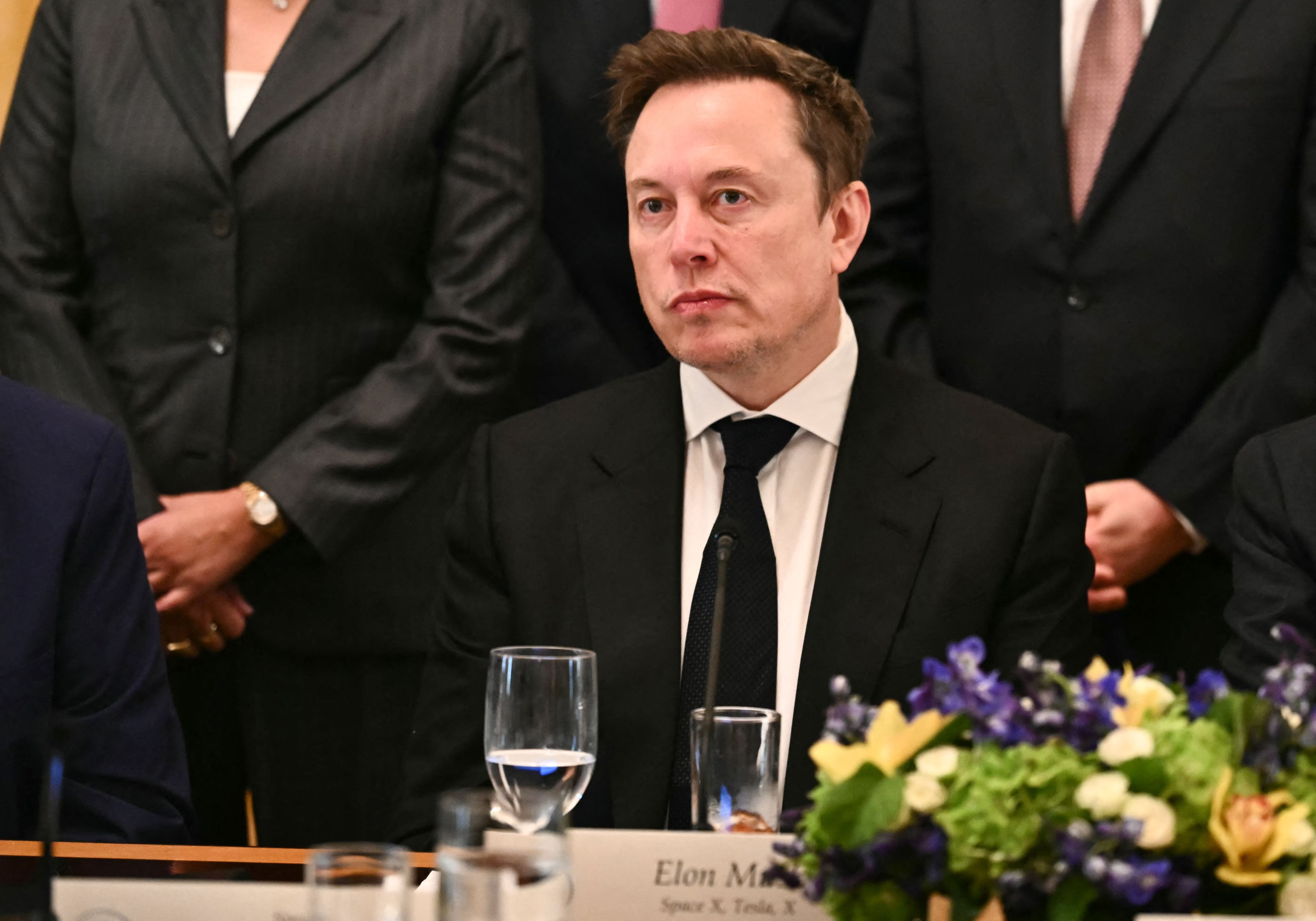 Ein Mann im Anzug sitzt an einem Konferenztisch mit Blumen und Wasserglas vor sich.