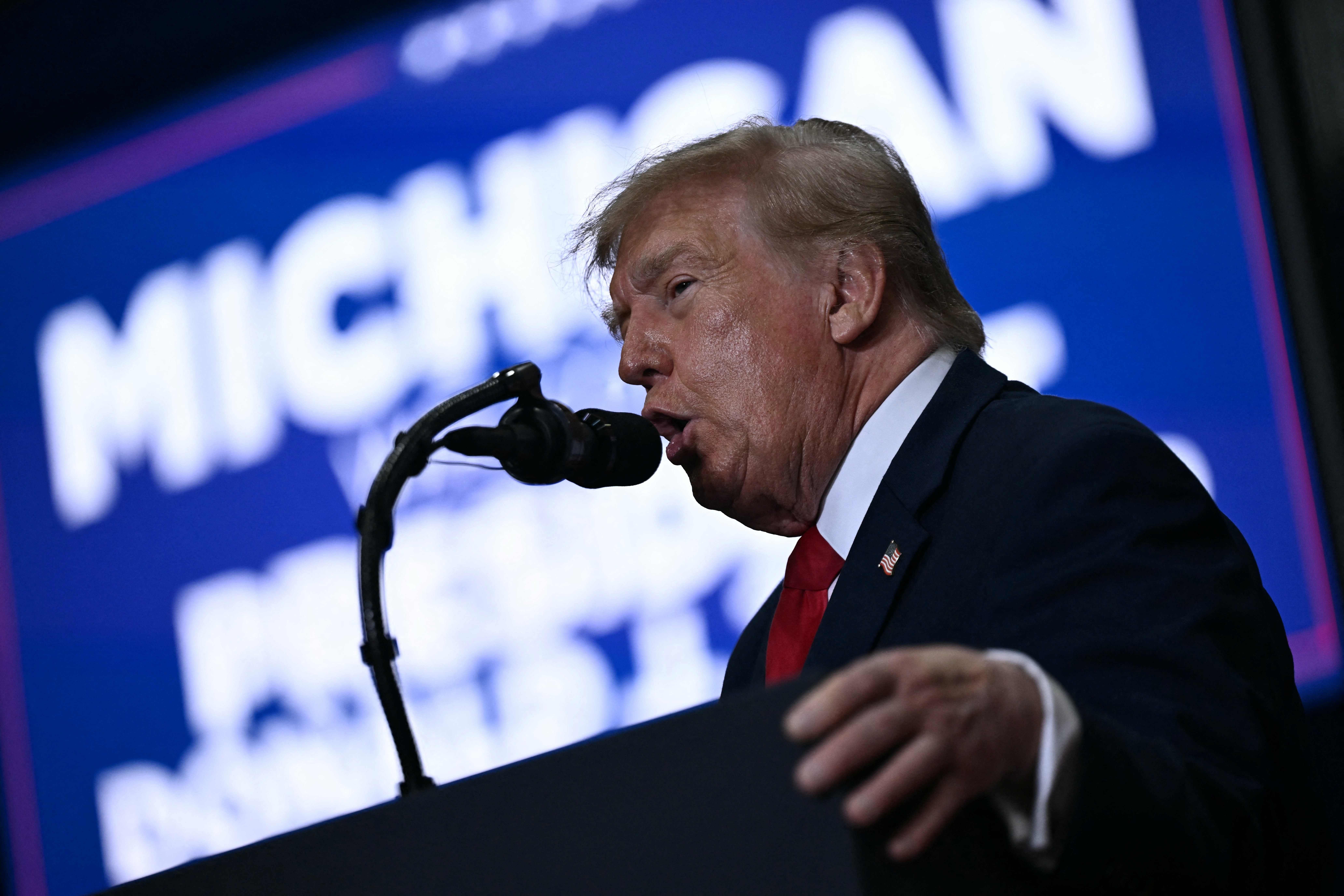 Ein Mann im Anzug spricht bei einer Veranstaltung an einem Mikrofon vor "MICHIGAN"-Hintergrund.