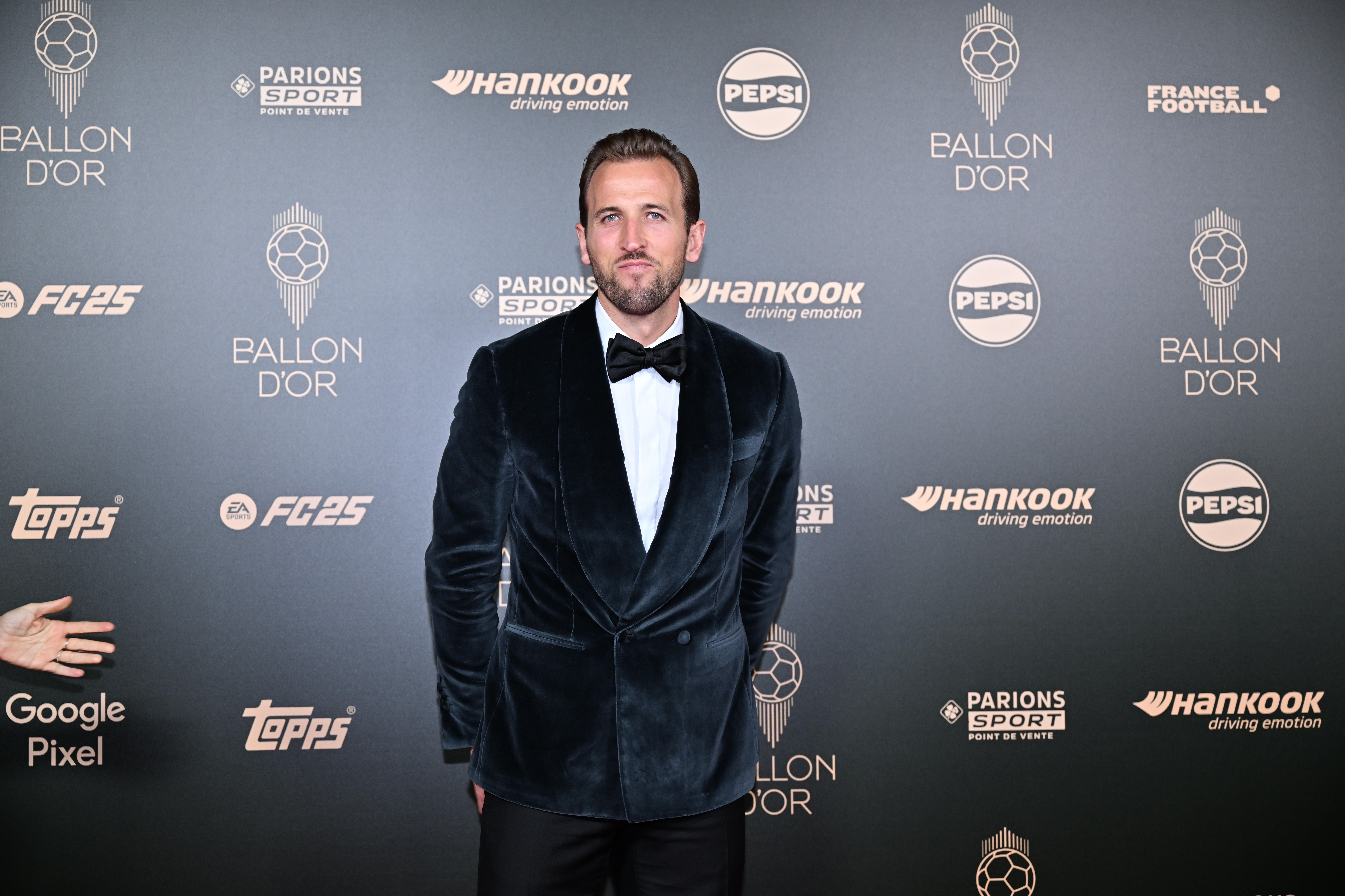 Ein Mann im Samtanzug posiert auf dem roten Teppich der Ballon d'Or-Gala vor Sponsorenwänden.