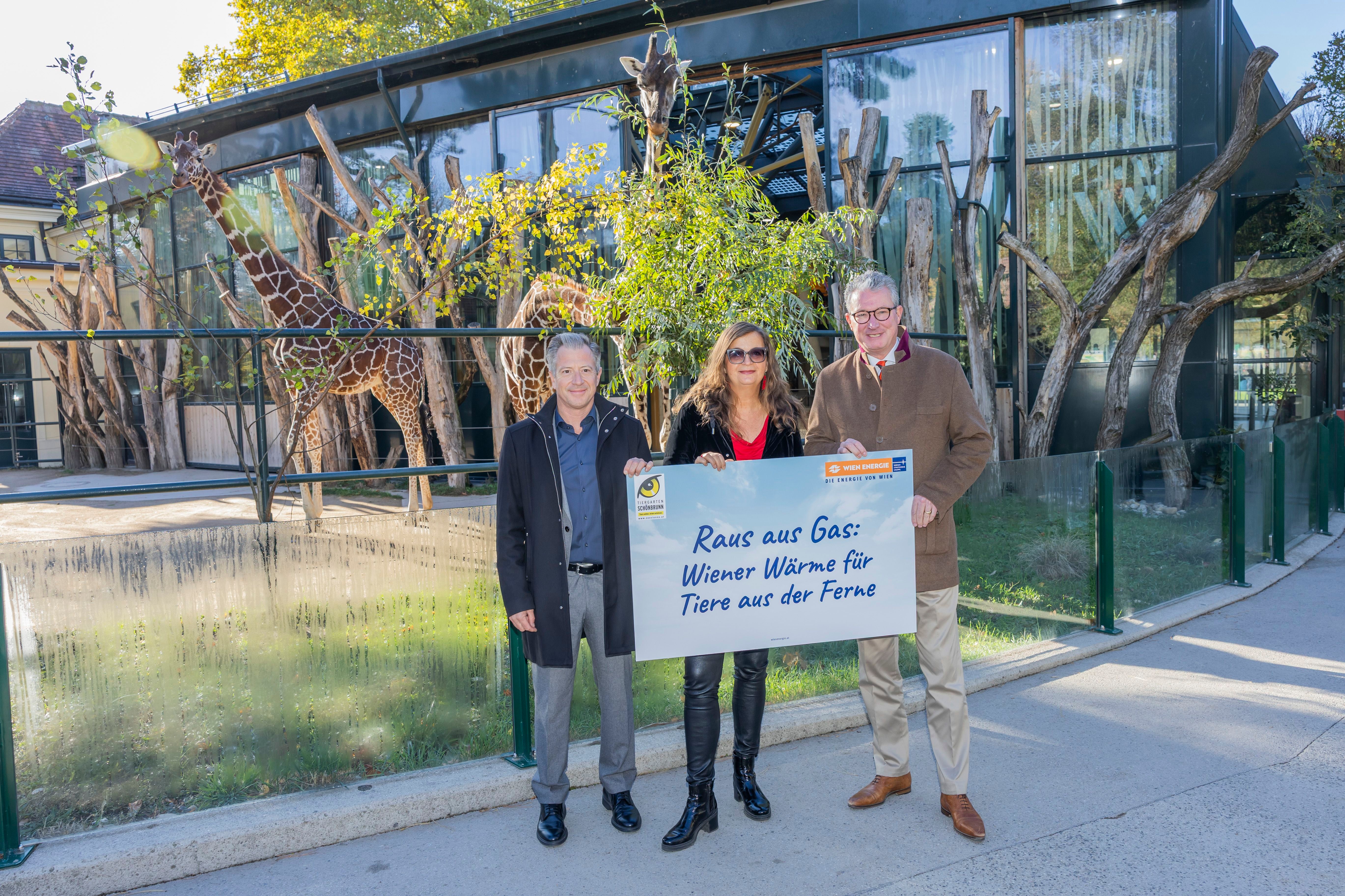 Drei Personen halten ein Schild vor einem Giraffengehege im Tiergarten Schönbrunn.