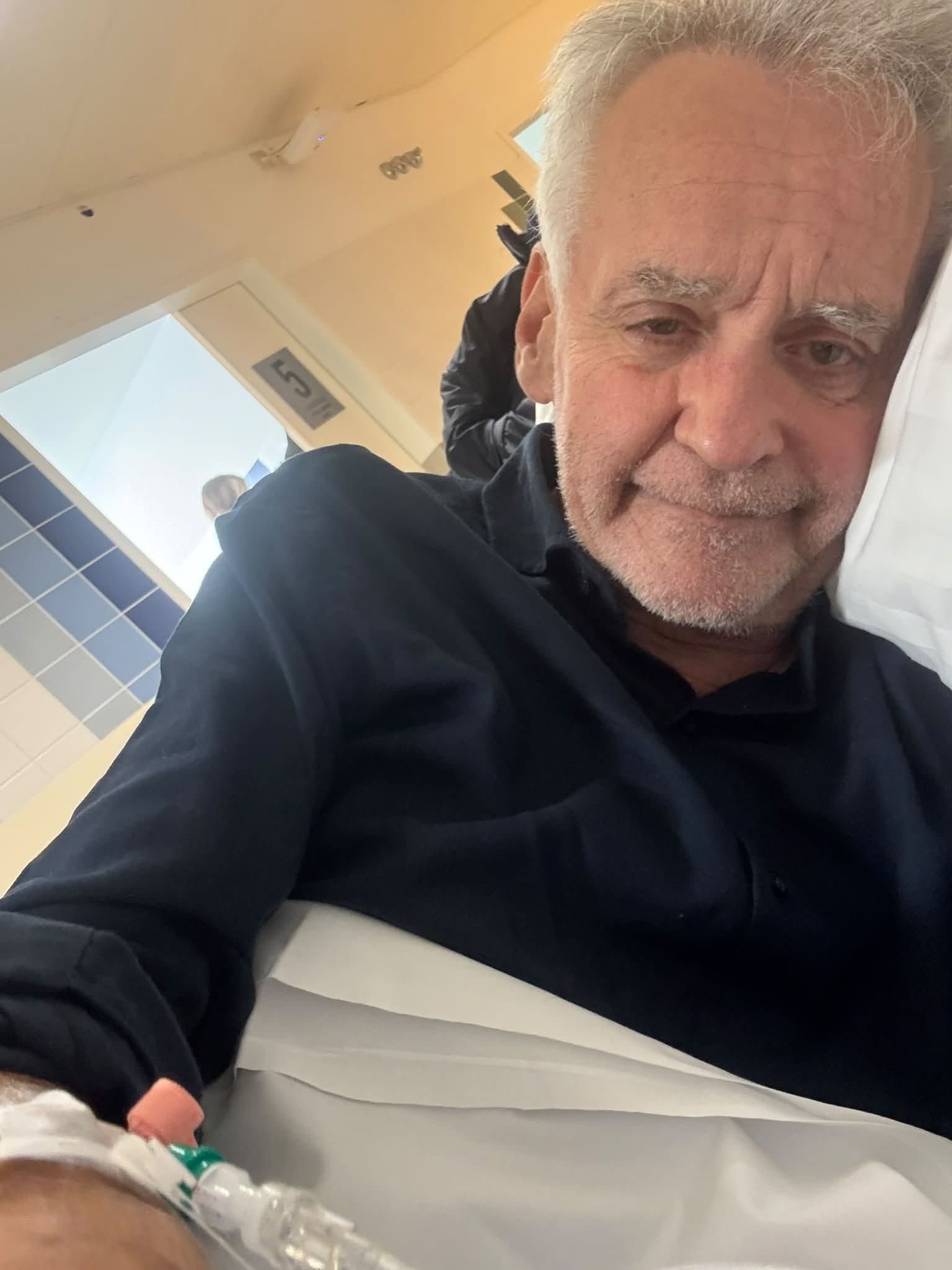 Älterer Mann liegt im Krankenhausbett mit Infusionsnadel im Arm und schaut in die Kamera.