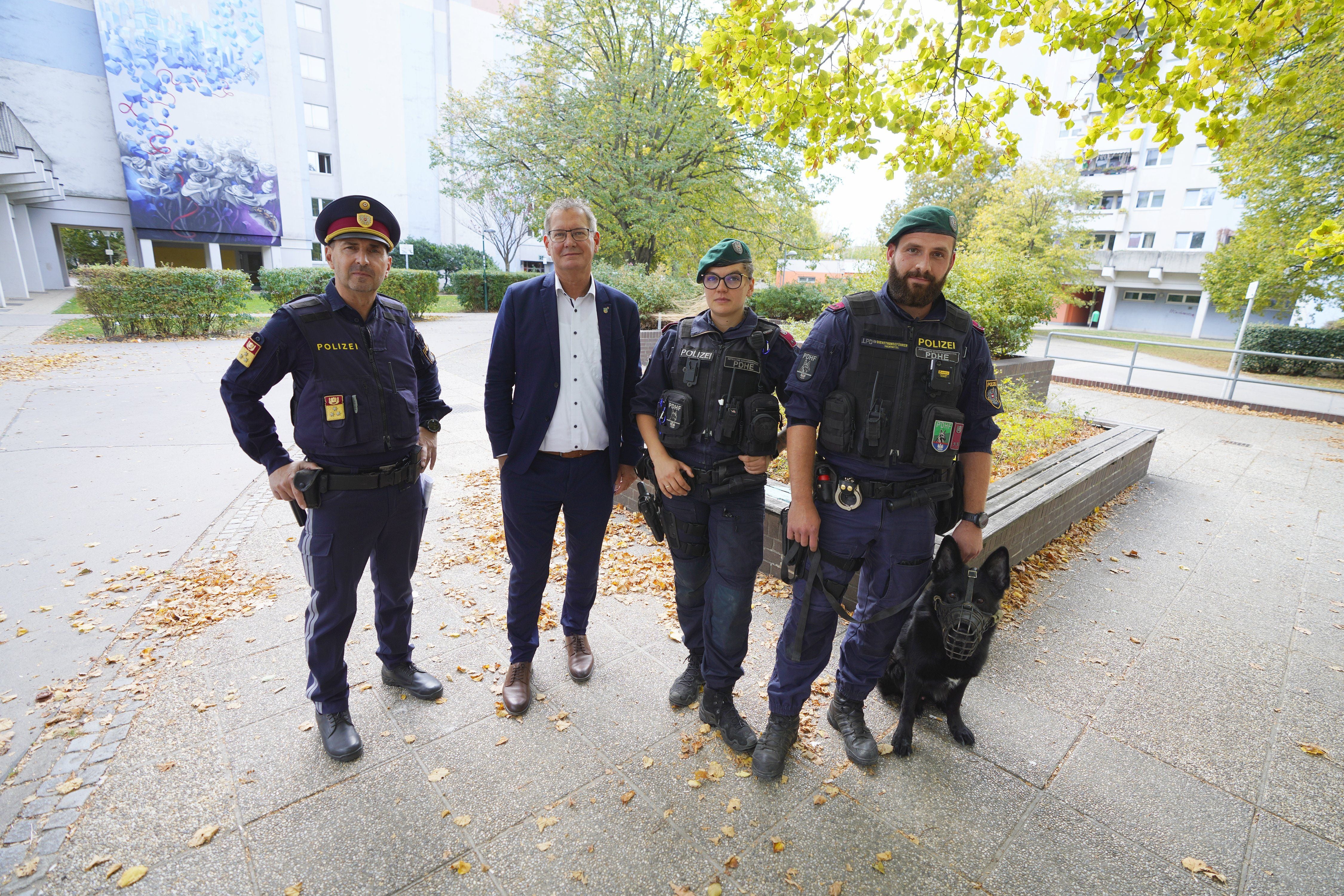 Vier Polizisten, einer mit Hund, und ein Mann in Zivil stehen im Freien vor Wohngebäuden.