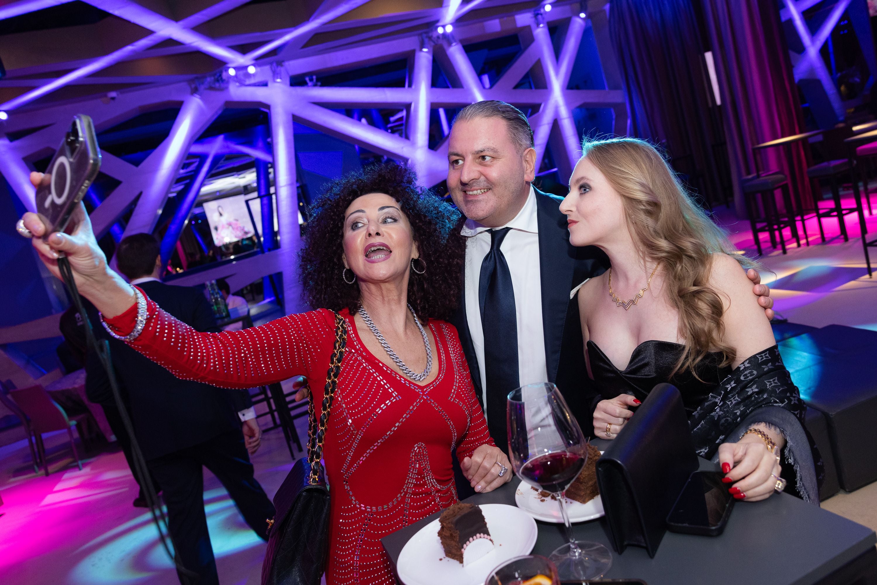 Drei elegant gekleidete Personen machen ein Selfie auf einer Party mit bunter Beleuchtung.
