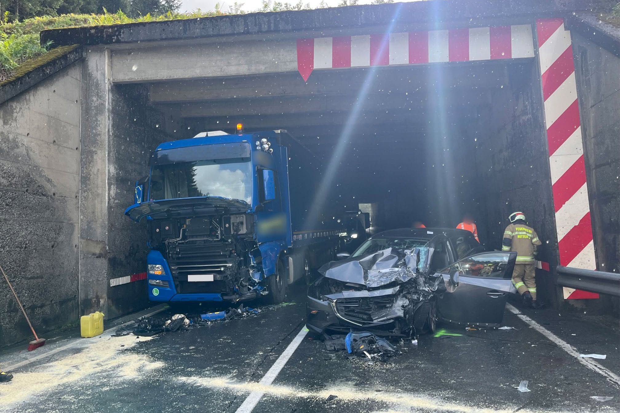 Unfall in einem Tunnel mit einem beschädigten blauen LKW, einem Auto und Einsatzkräften vor Ort.