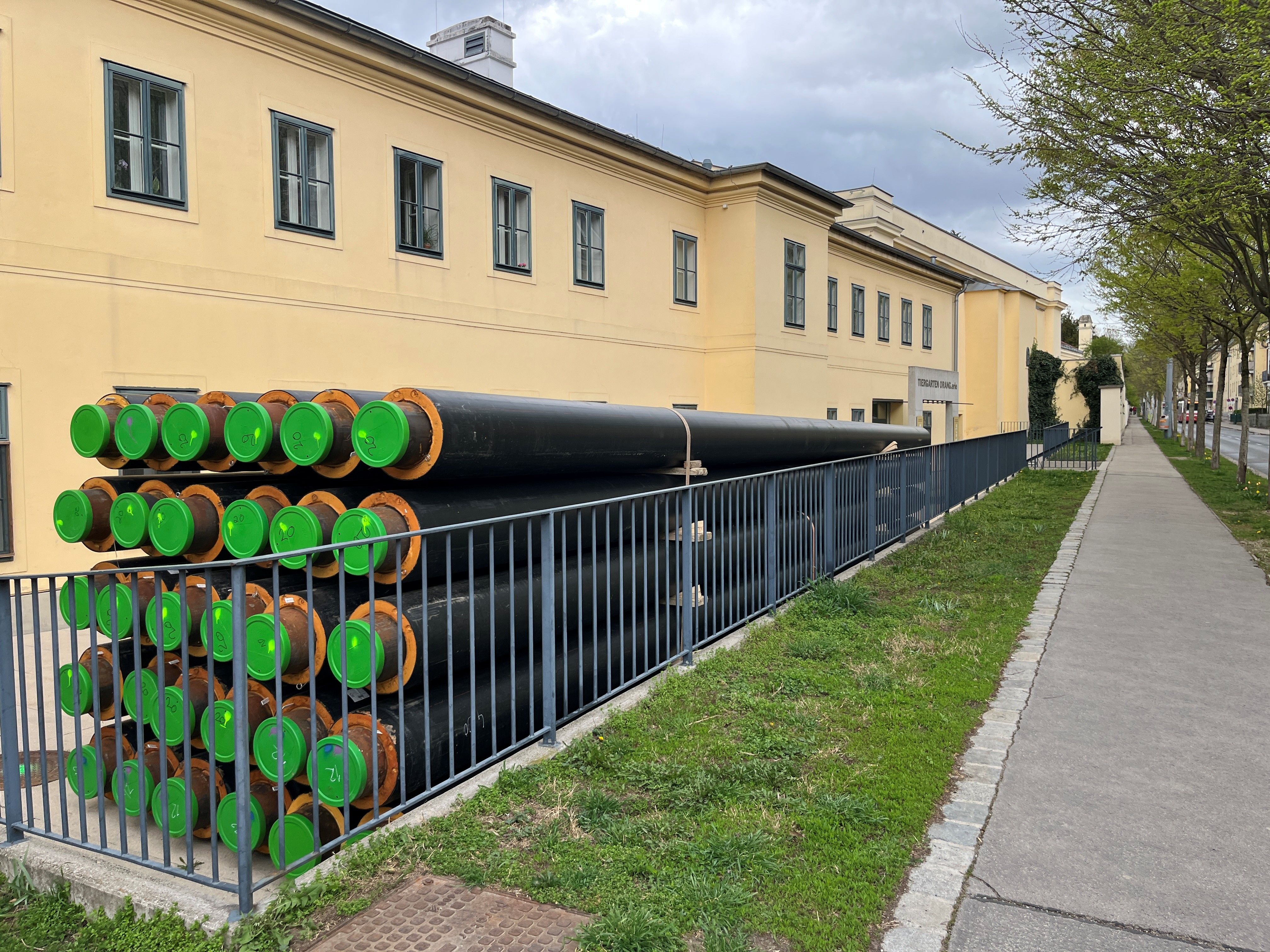 Große schwarze Fernwärmerohre mit grünen Endkappen liegen gestapelt neben einem gelben Gebäude.