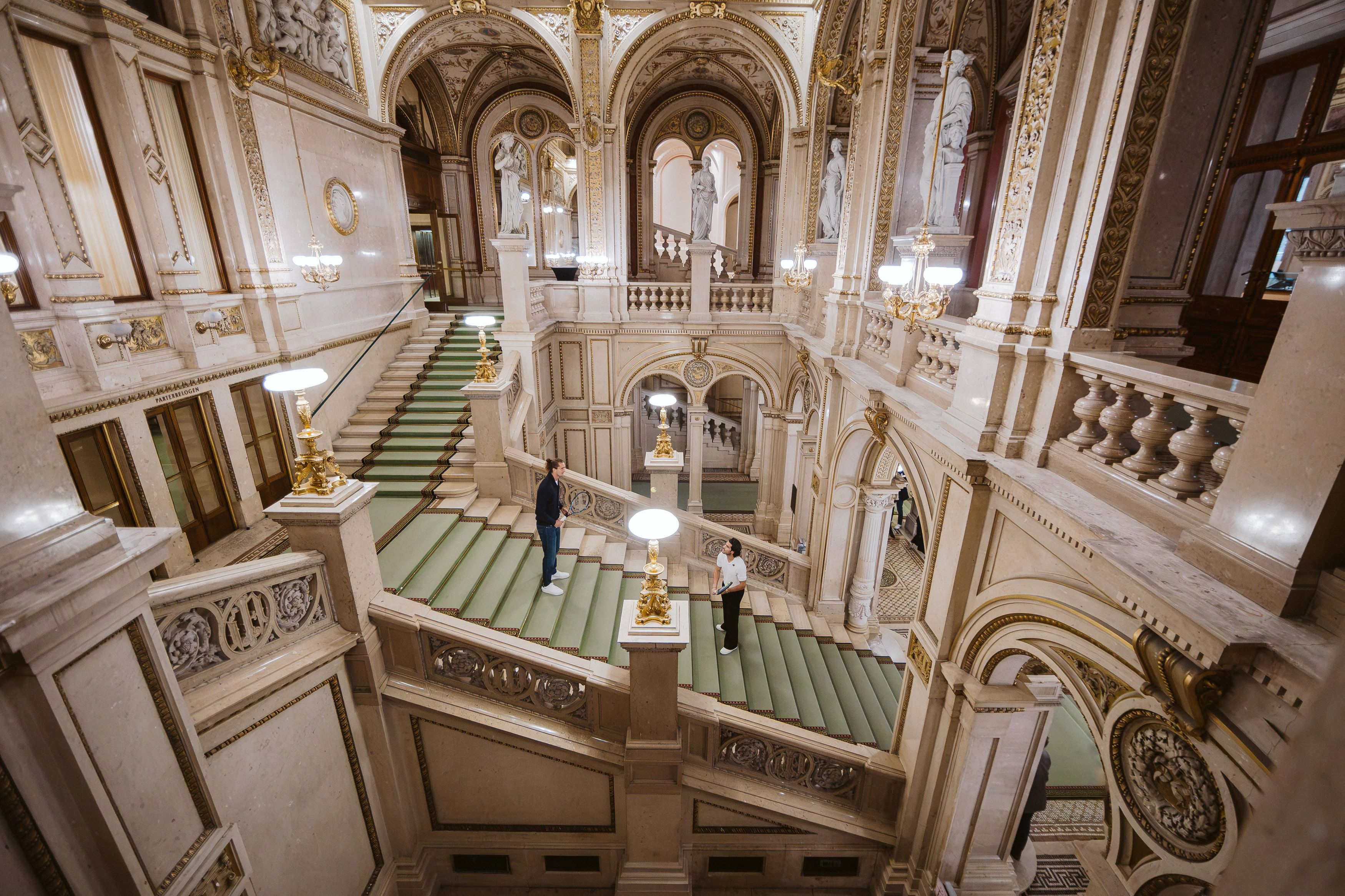 Zwei Personen stehen auf einer prunkvollen, breiten Marmortreppe in einem historischen Gebäude.