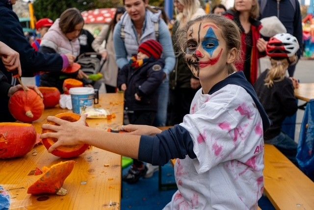 Kind mit Clown-Gesichtsbemalung schnitzt an einem Halloween-Tisch einen Kürbis.