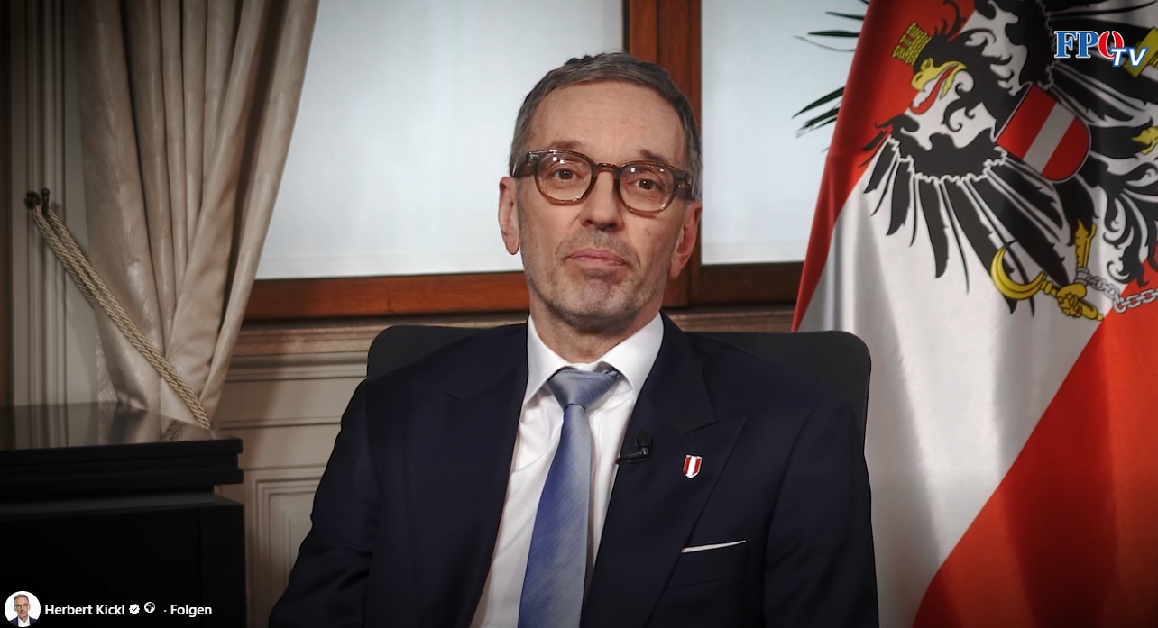 Ein Mann im Anzug hält eine Ansprache, im Hintergrund eine österreichische Flagge.