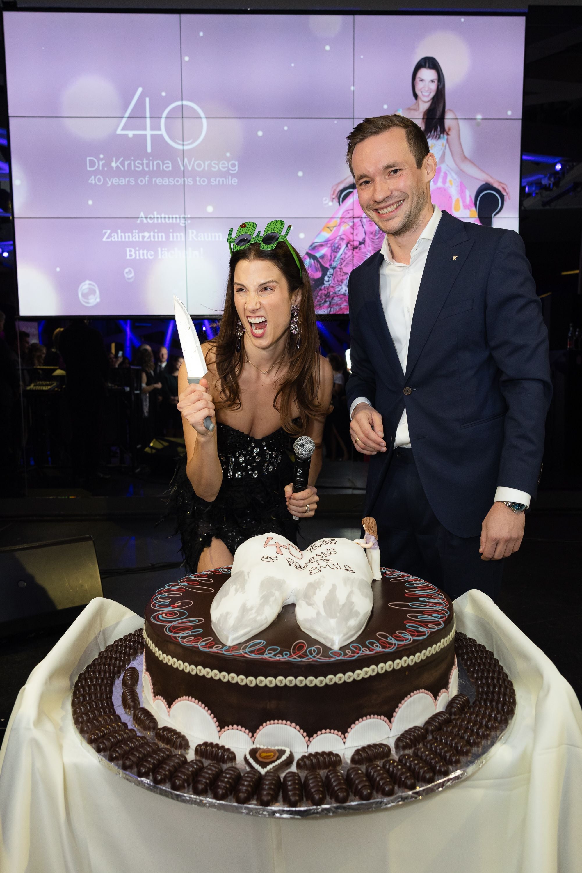 Frau mit Geburtstagsbrille posiert mit Mann und Messer vor großer Torte mit Zahn-Design.