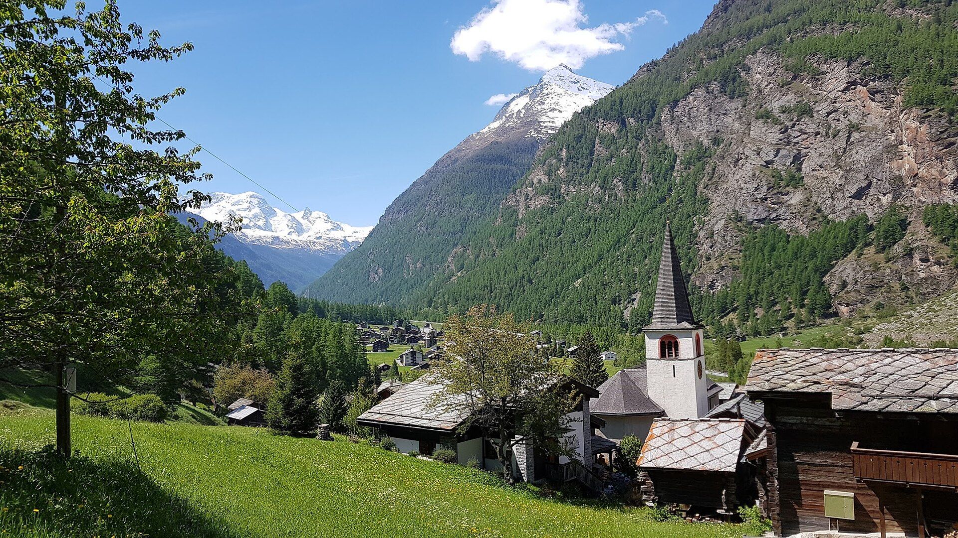 Bergdorf Randa mit Kirche, Wiesen, Holzhäusern und schneebedeckten Alpen im Hintergrund.