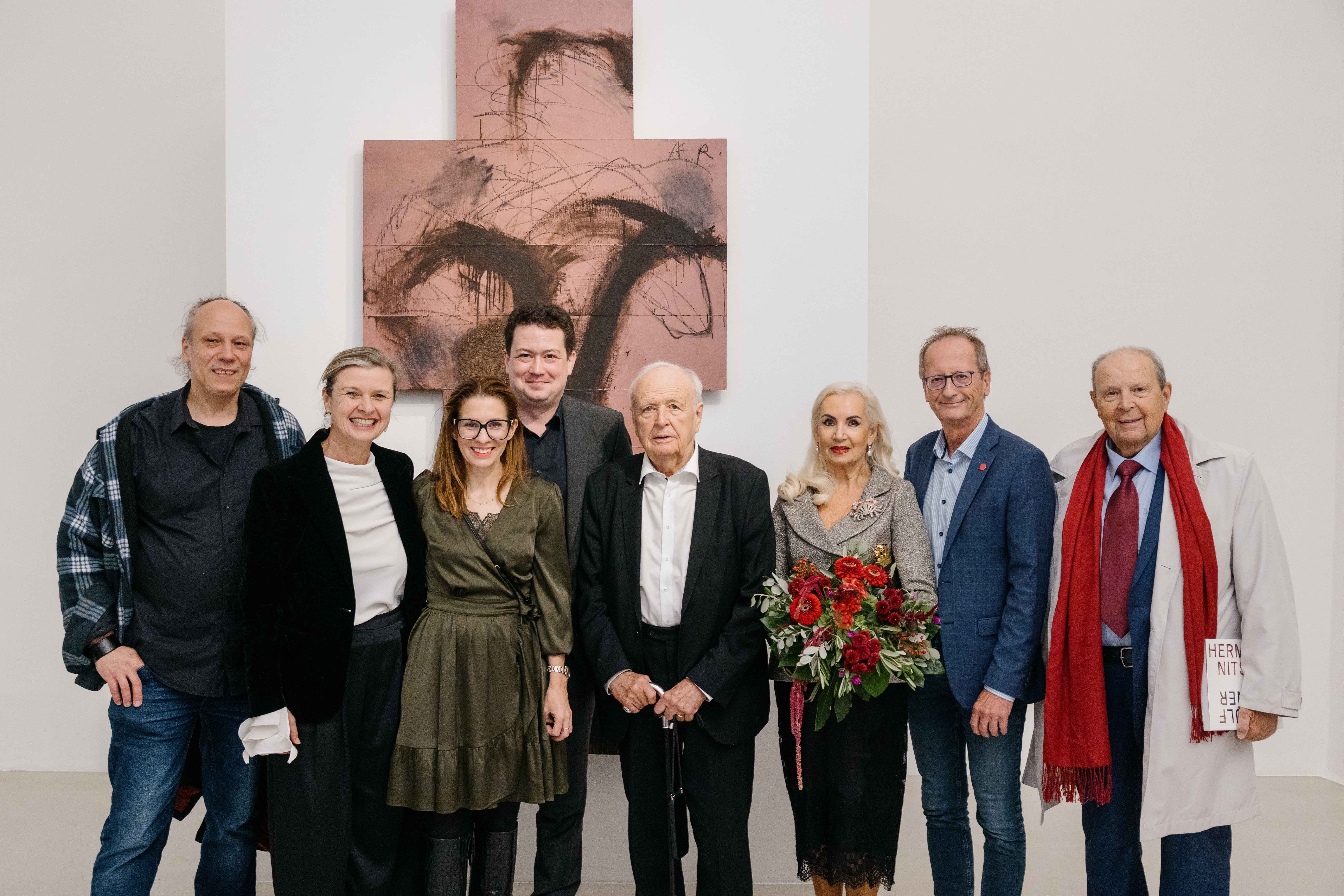 Acht Personen stehen vor abstrakter Kunst, eine Frau hält einen Blumenstrauß.