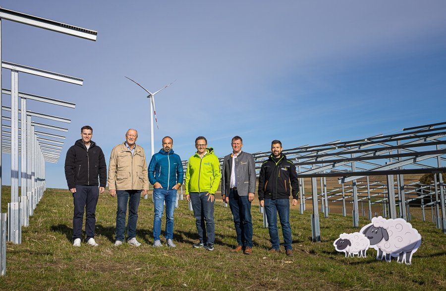 Sechs Männer stehen auf einer Wiese zwischen Metallgestellen, im Hintergrund Windrad und Schafzeichnungen.