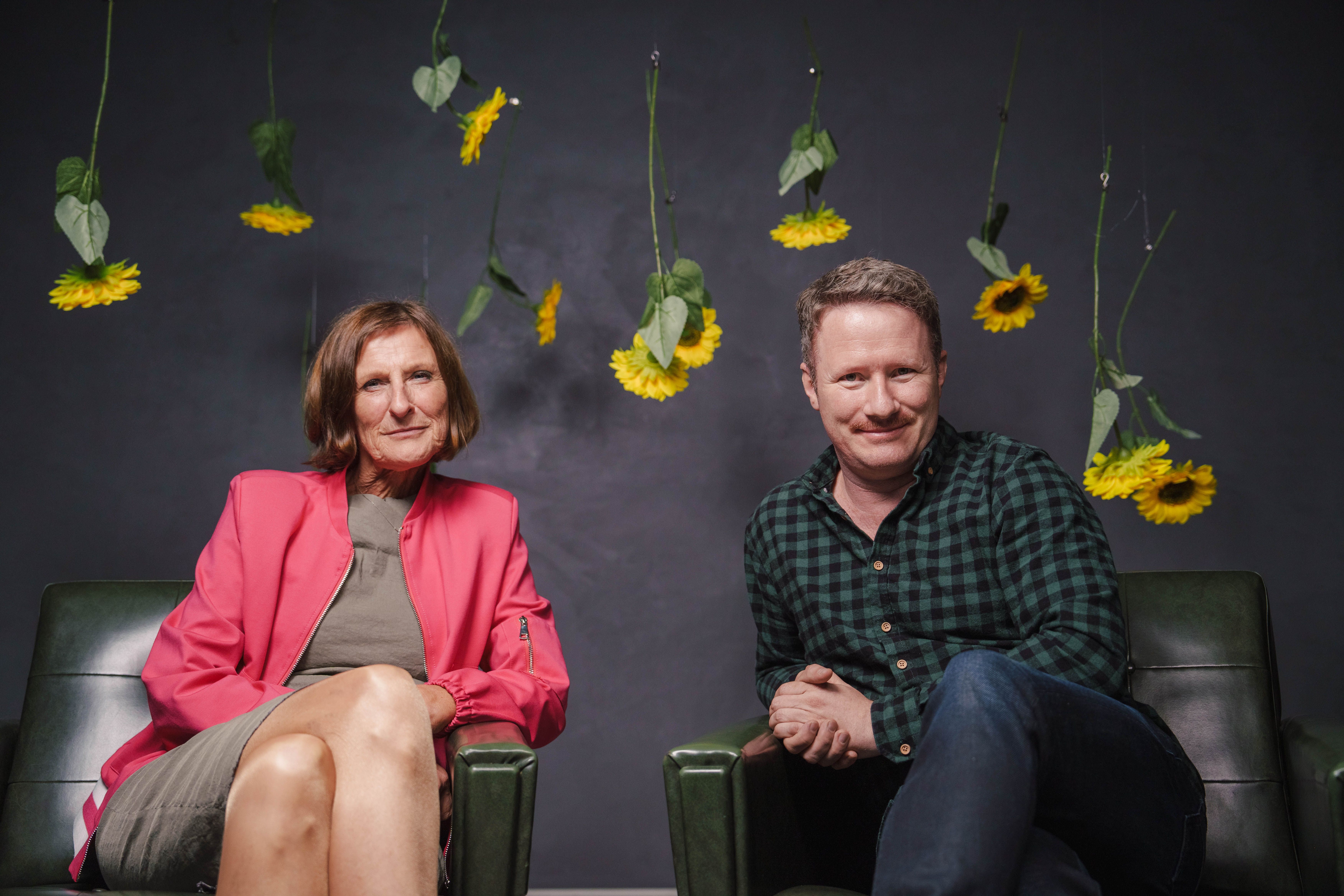 Zwei Personen sitzen vor hängenden Sonnenblumen in einem dunklen Raum.