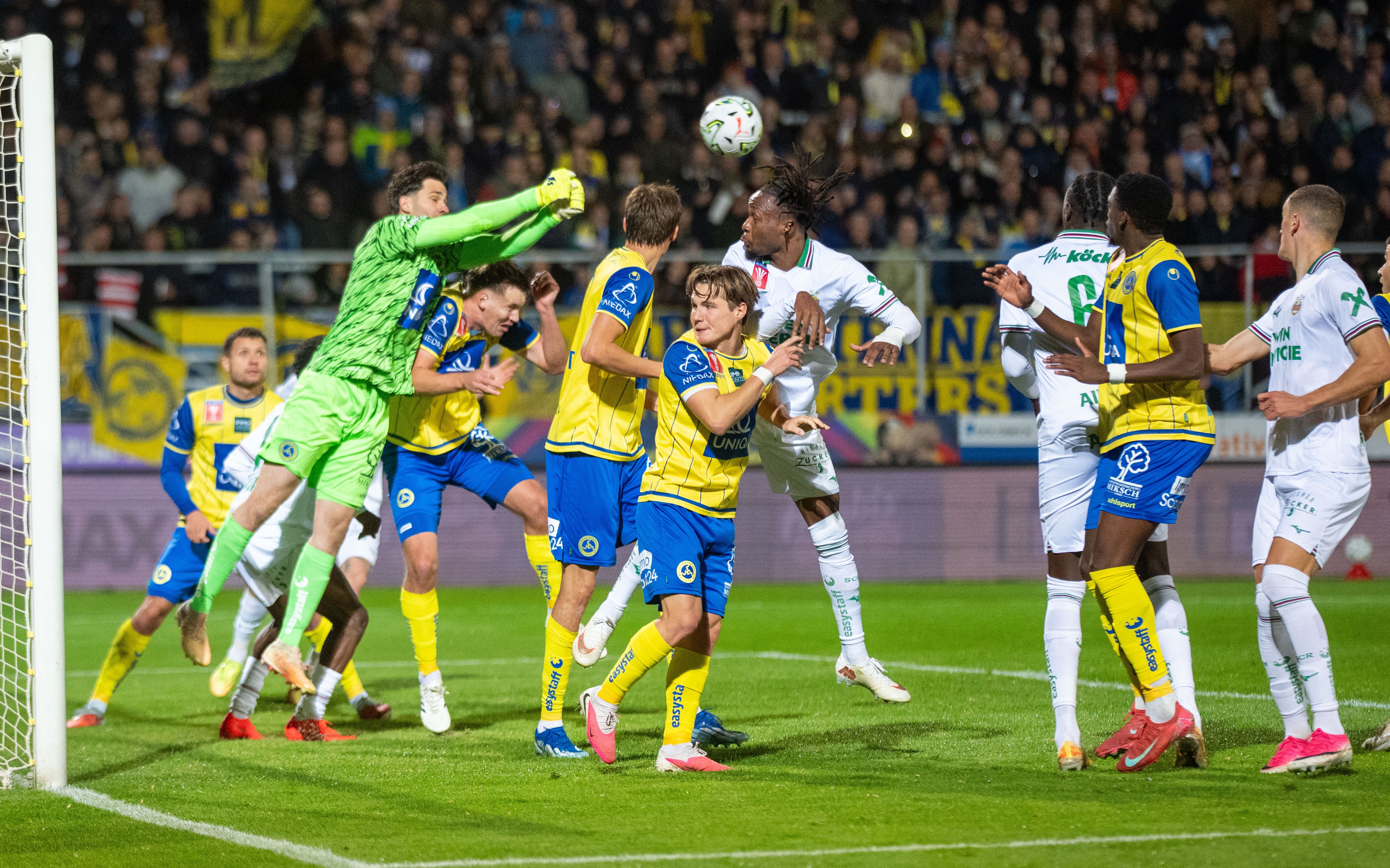 Fußballspiel: Torwart springt im Strafraum, mehrere Spieler kämpfen um den Ball.