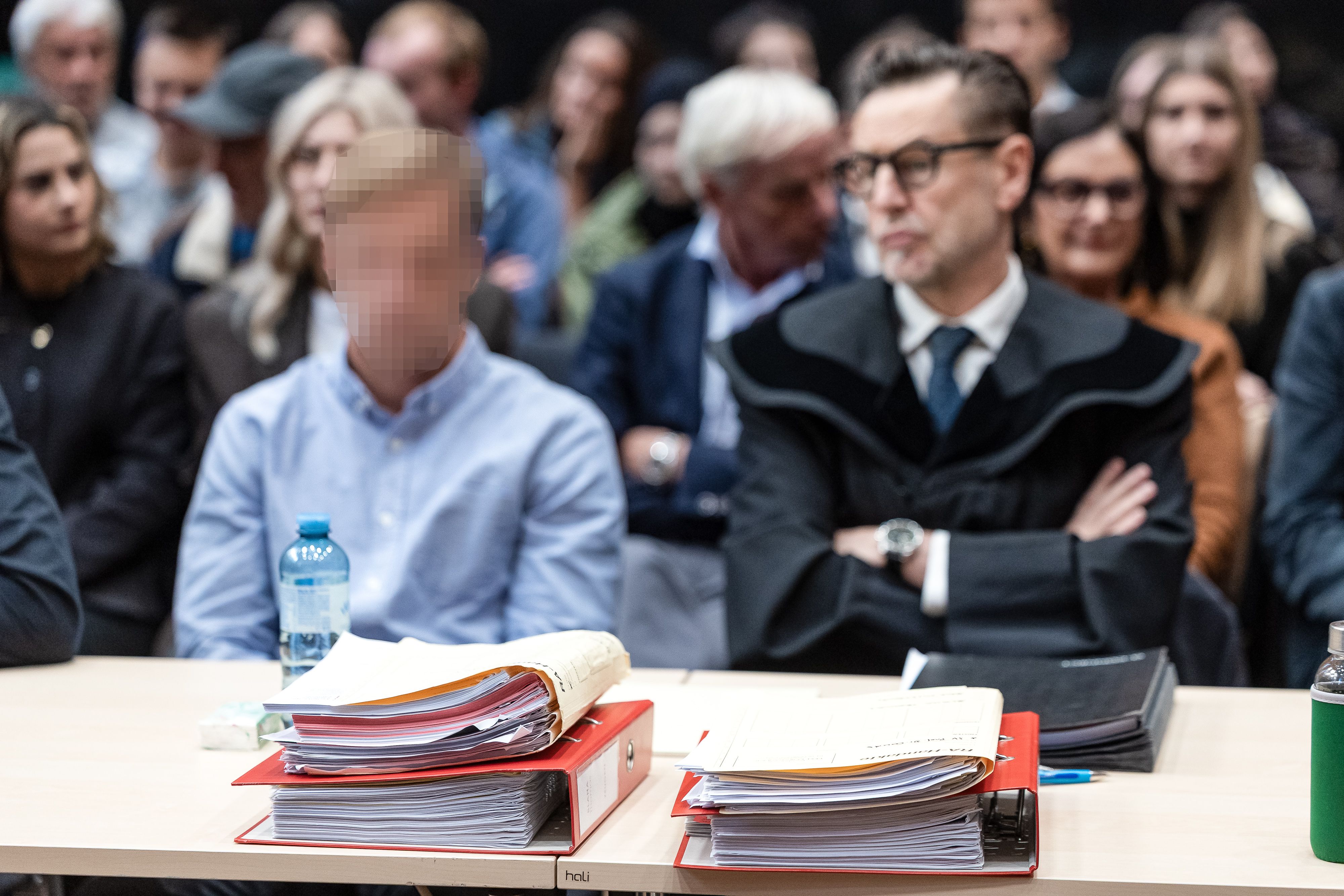 Ein Angeklagter und sein Anwalt sitzen vor Aktenordnern im Gerichtssaal, im Hintergrund Zuschauer.
