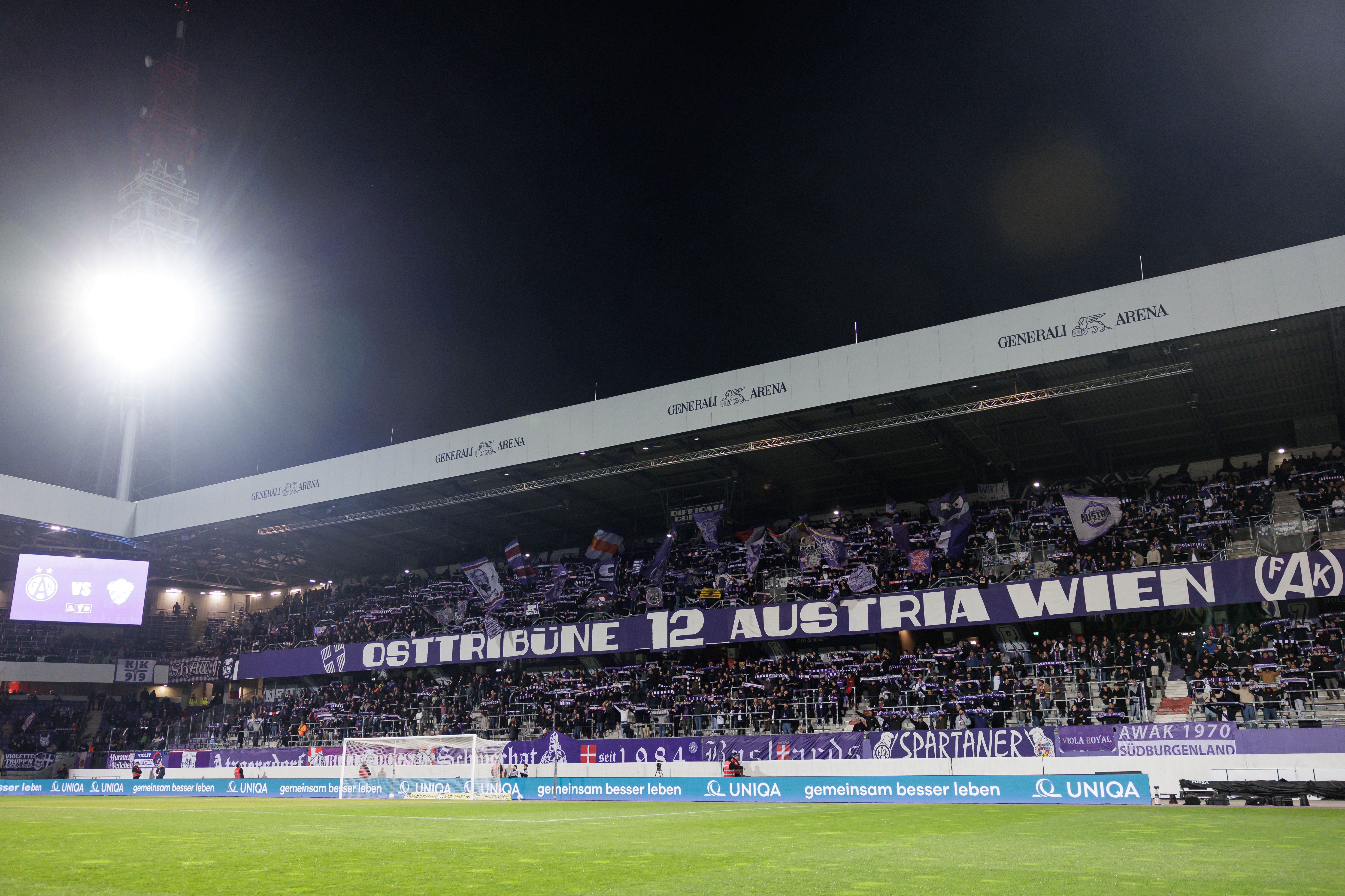 Die gut gefüllte Osttribüne der Generali Arena mit dem großen Banner Osttribüne 12 Austria Wien unter Flutlicht während eines Heimspiels am Verteilerkreis.