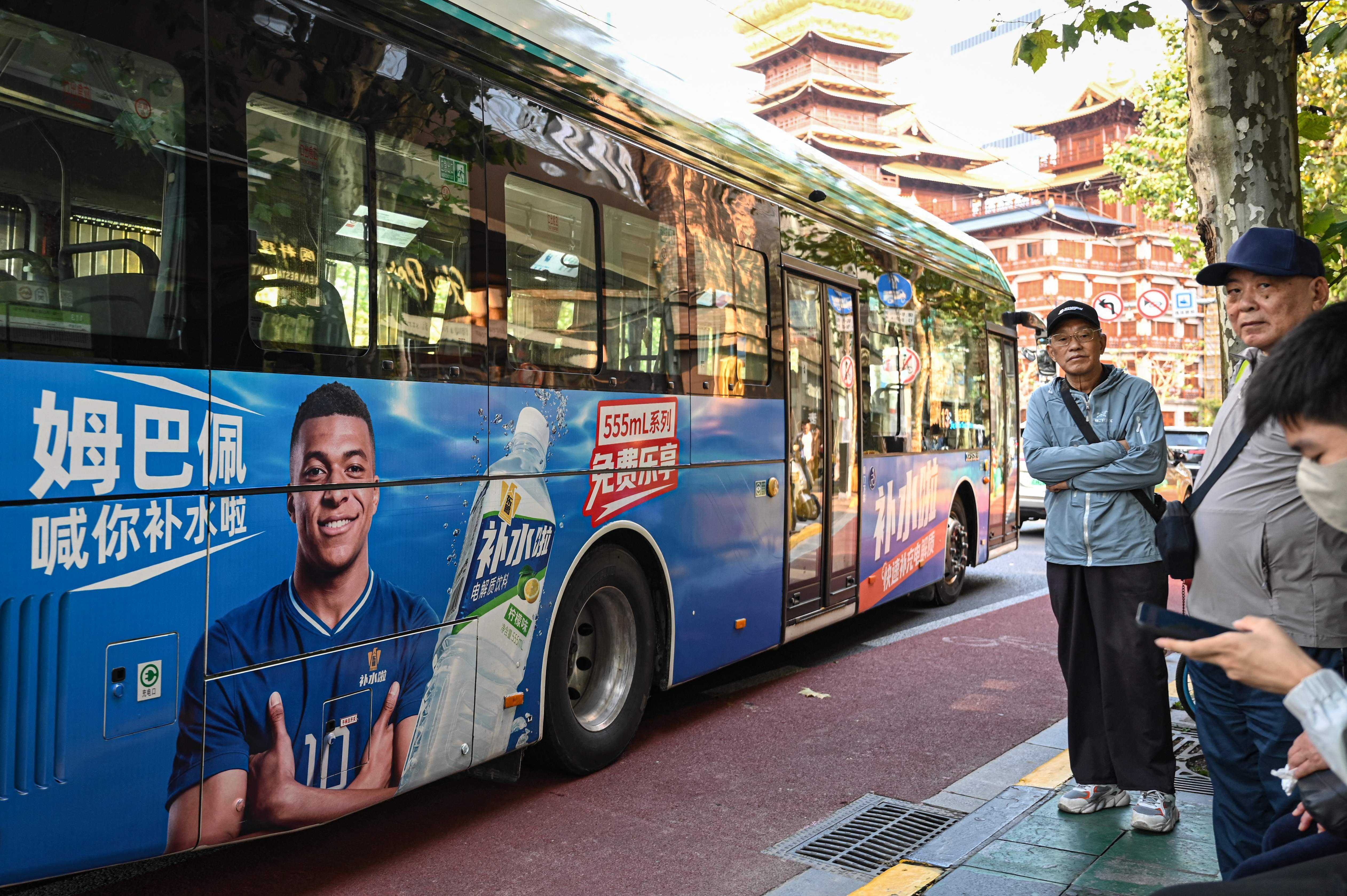Menschen stehen an einer Bushaltestelle in Shanghai neben einem Bus mit Werbung von Kylian Mbappé.