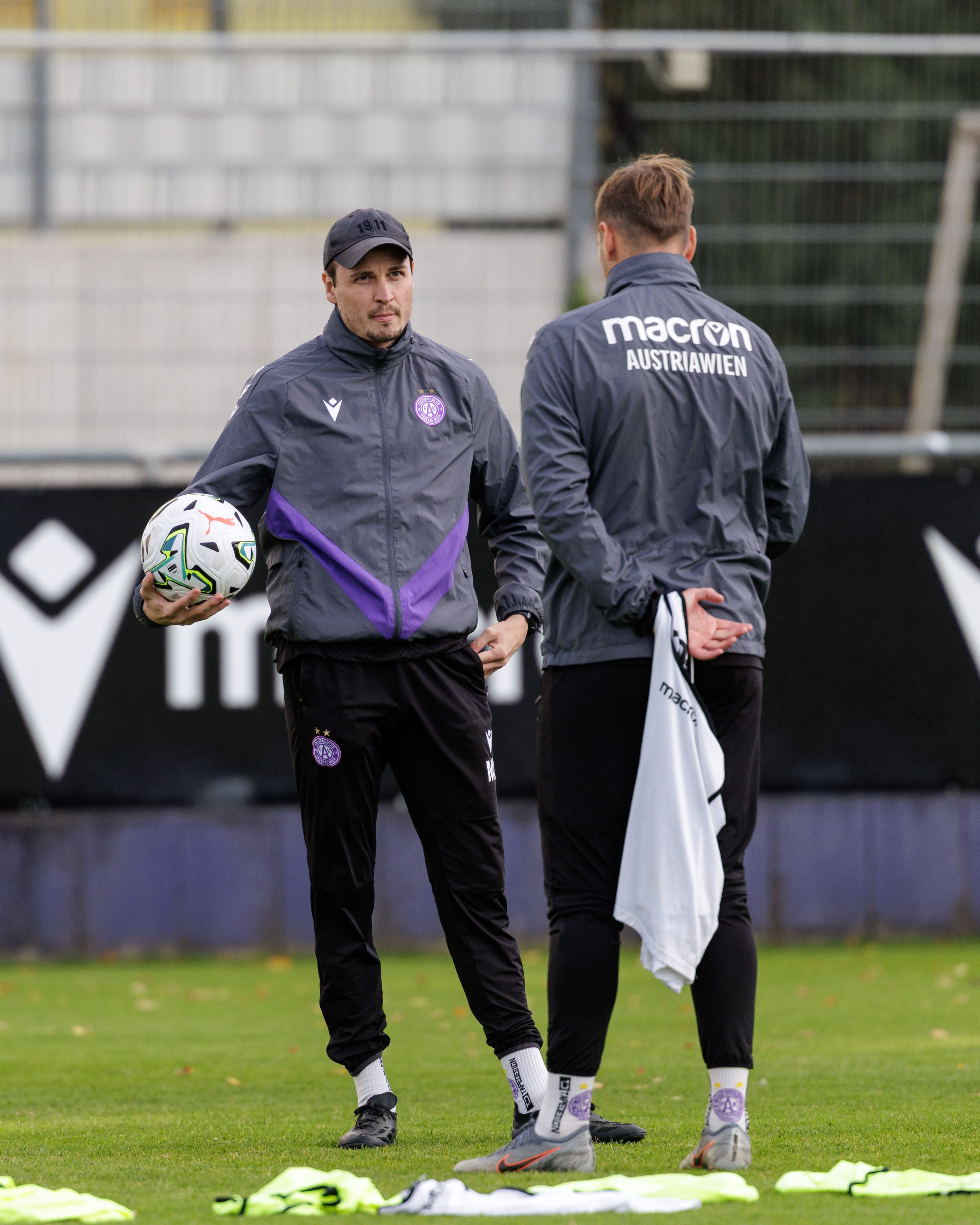 Zwei Fußballtrainer im Trainingsanzug unterhalten sich auf dem Spielfeld, einer hält einen Ball.