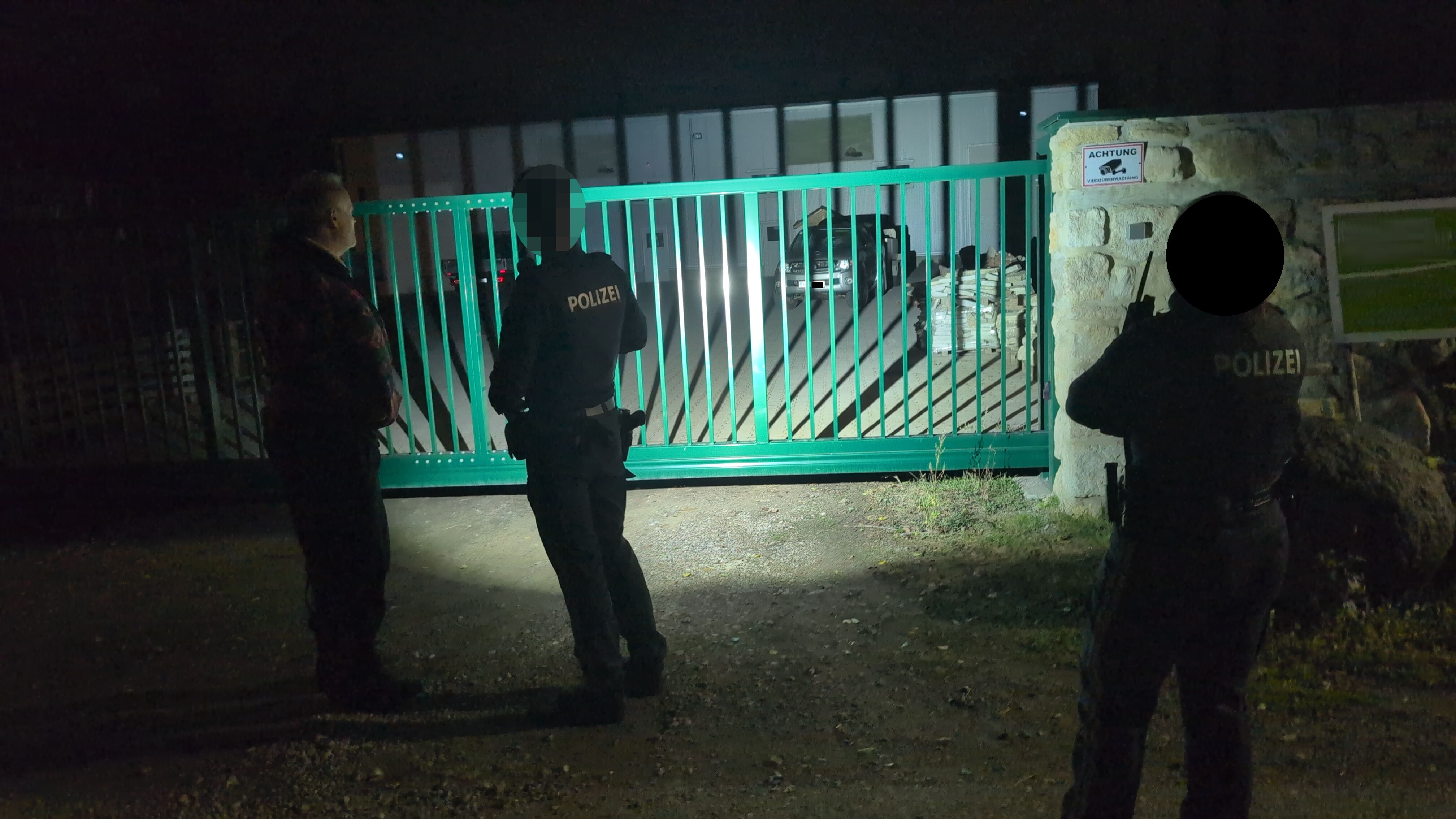 Drei Personen, darunter zwei Polizisten, stehen nachts vor einem verschlossenen Metalltor.
