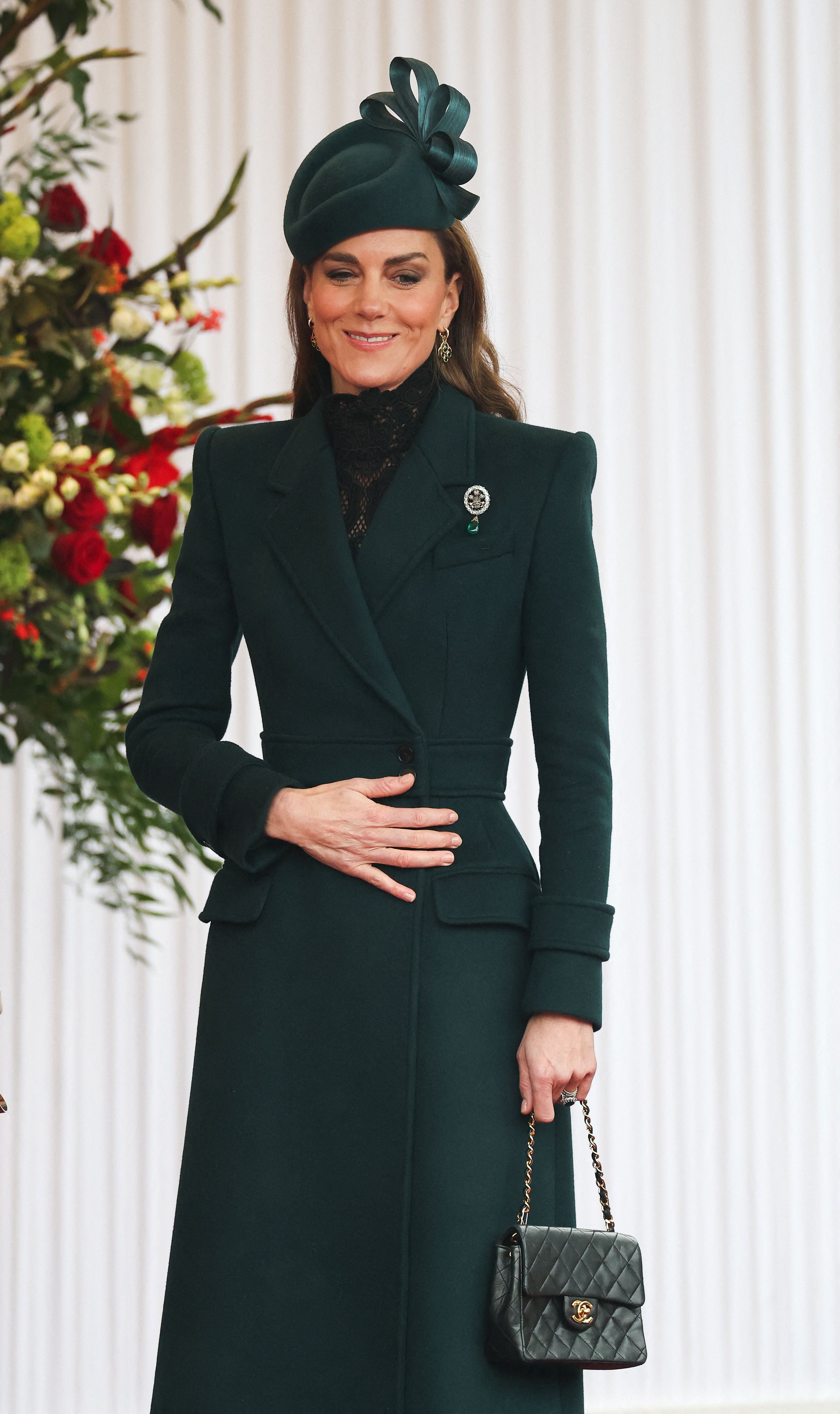Eine Frau in elegantem, dunkelgrünem Mantel und Hut hält eine schwarze Handtasche.