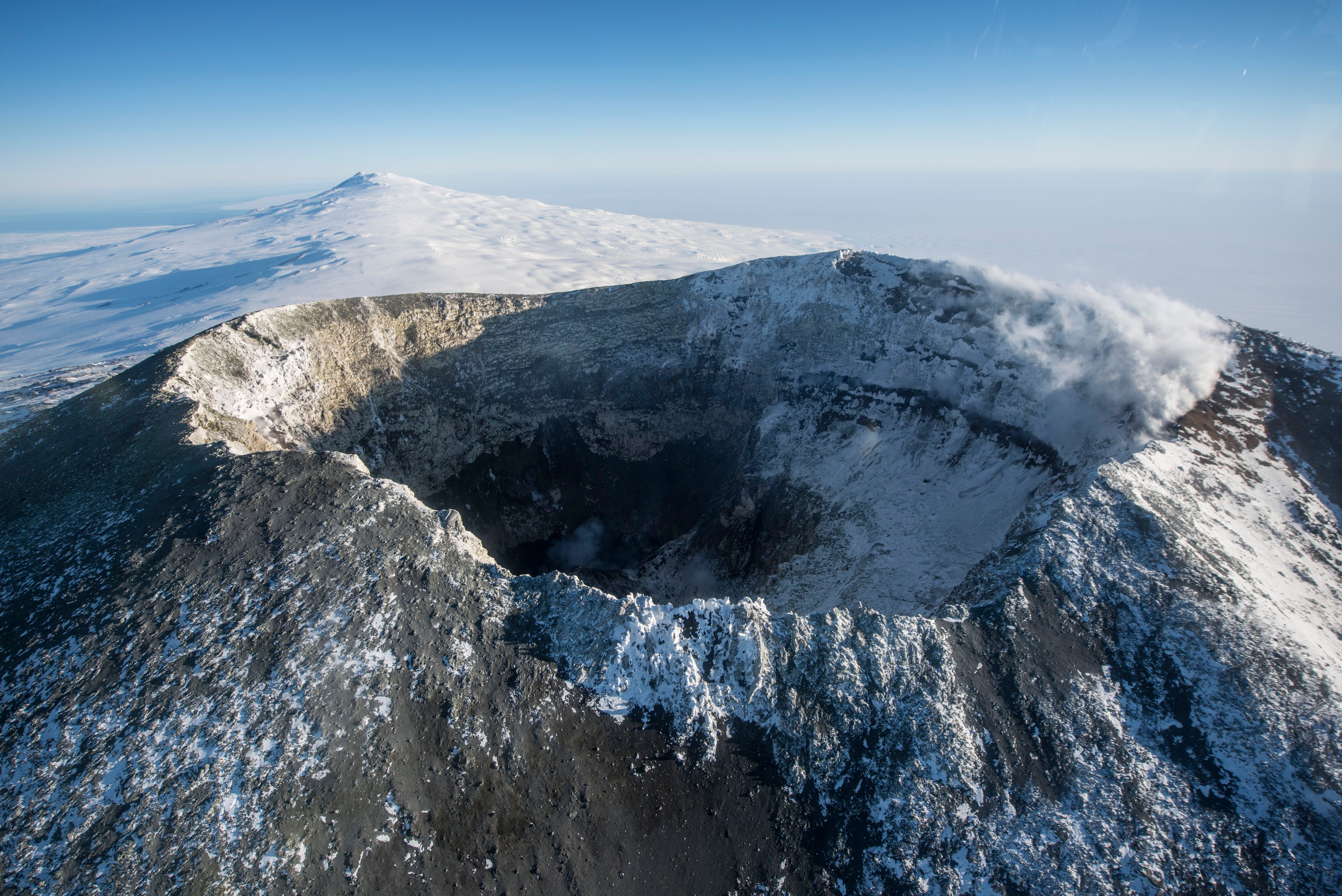 Luftaufnahme des schneebedeckten Vulkans Mount Erebus mit rauchendem Krater in eisiger Landschaft.