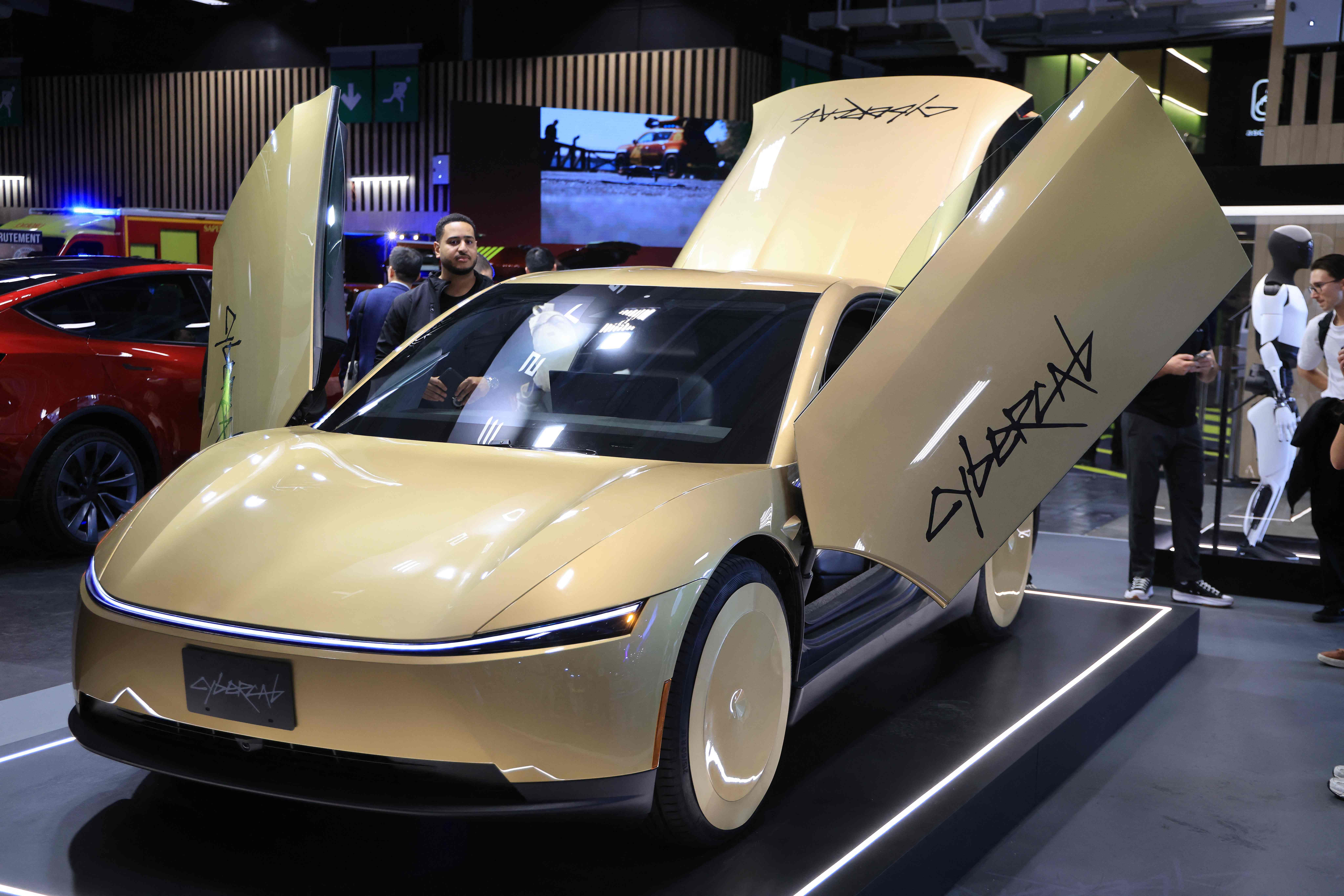 Goldener Tesla Cybercab mit Flügeltüren auf einer Automesse; futuristisches, autonomes Elektroauto.