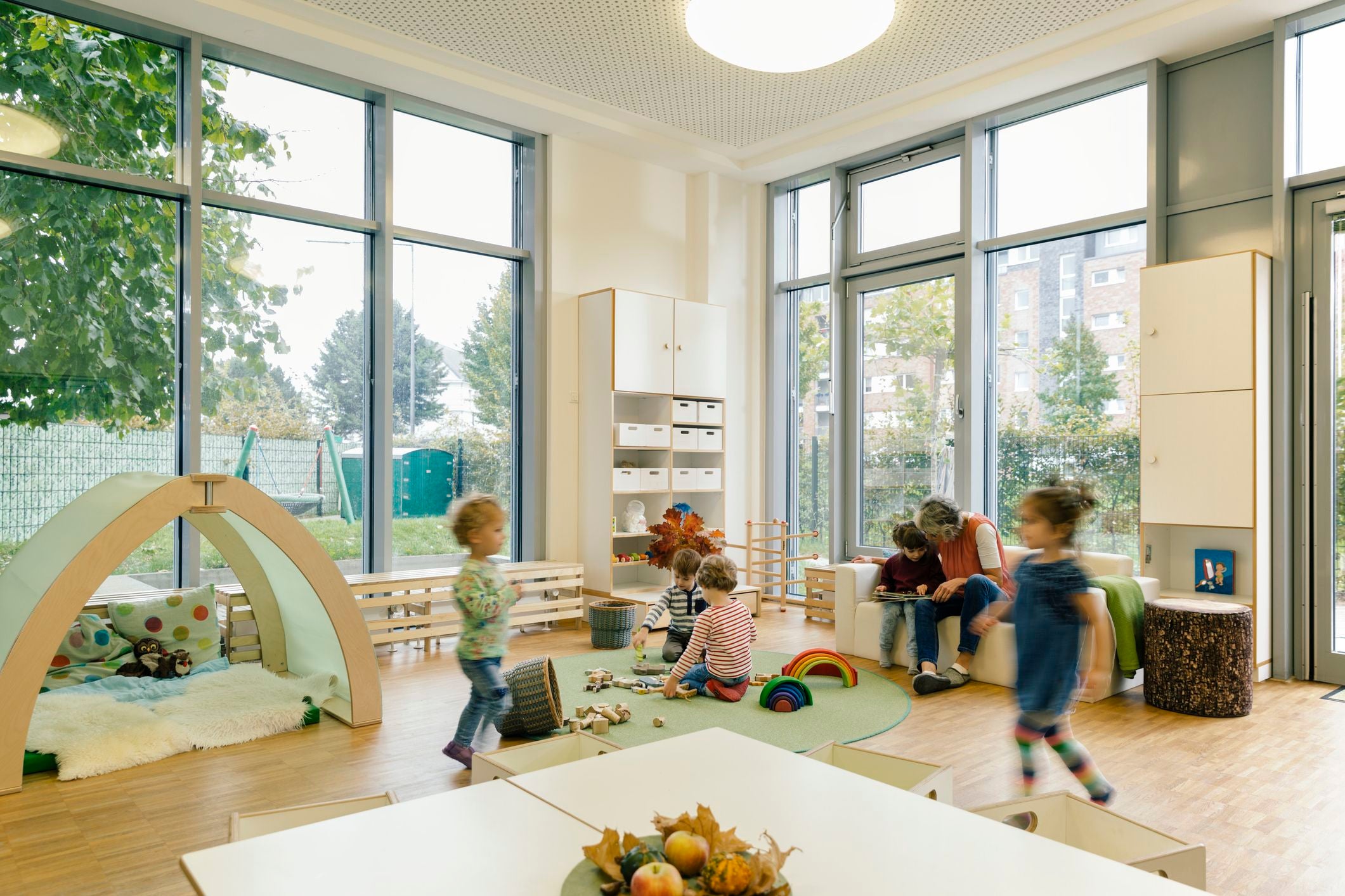 Kinder spielen in einem hellen Kindergartenraum mit großen Fenstern und Spielzeug auf dem Boden.