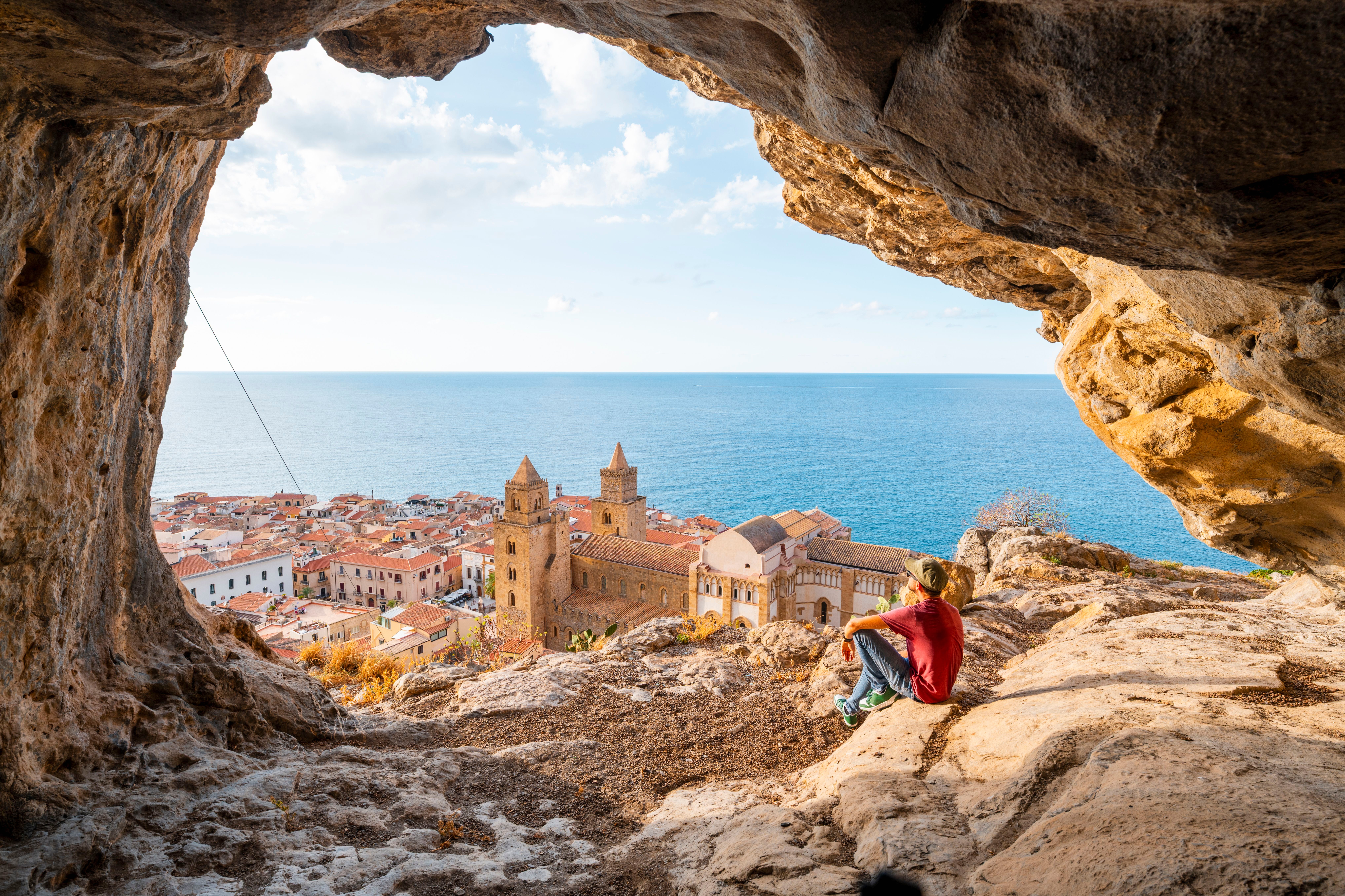 Mann sitzt in einer Felshöhle mit Blick auf eine Küstenstadt und das Meer.