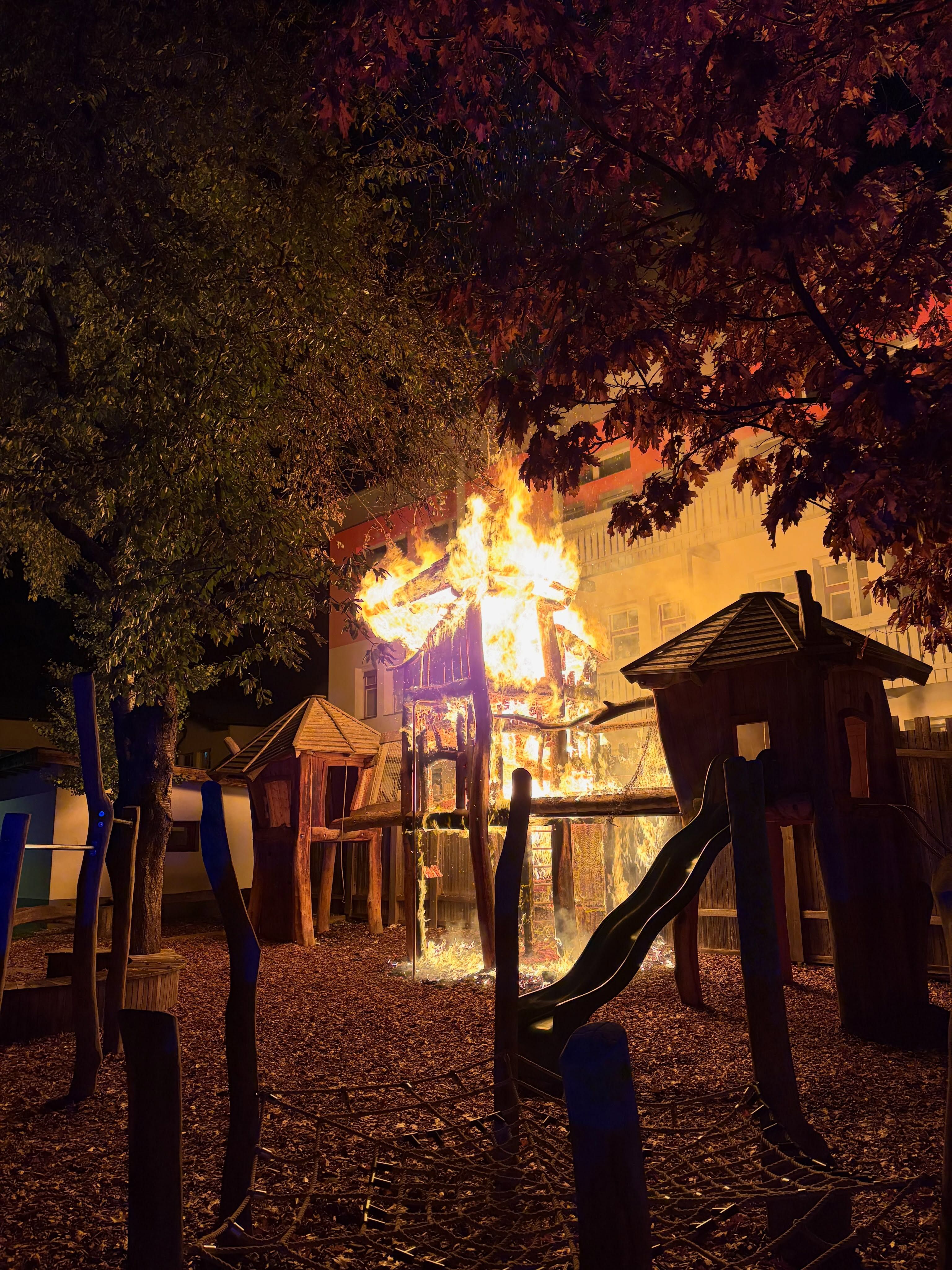 Großes Klettergerüst auf einem Spielplatz steht nachts in Flammen, umgeben von Bäumen.