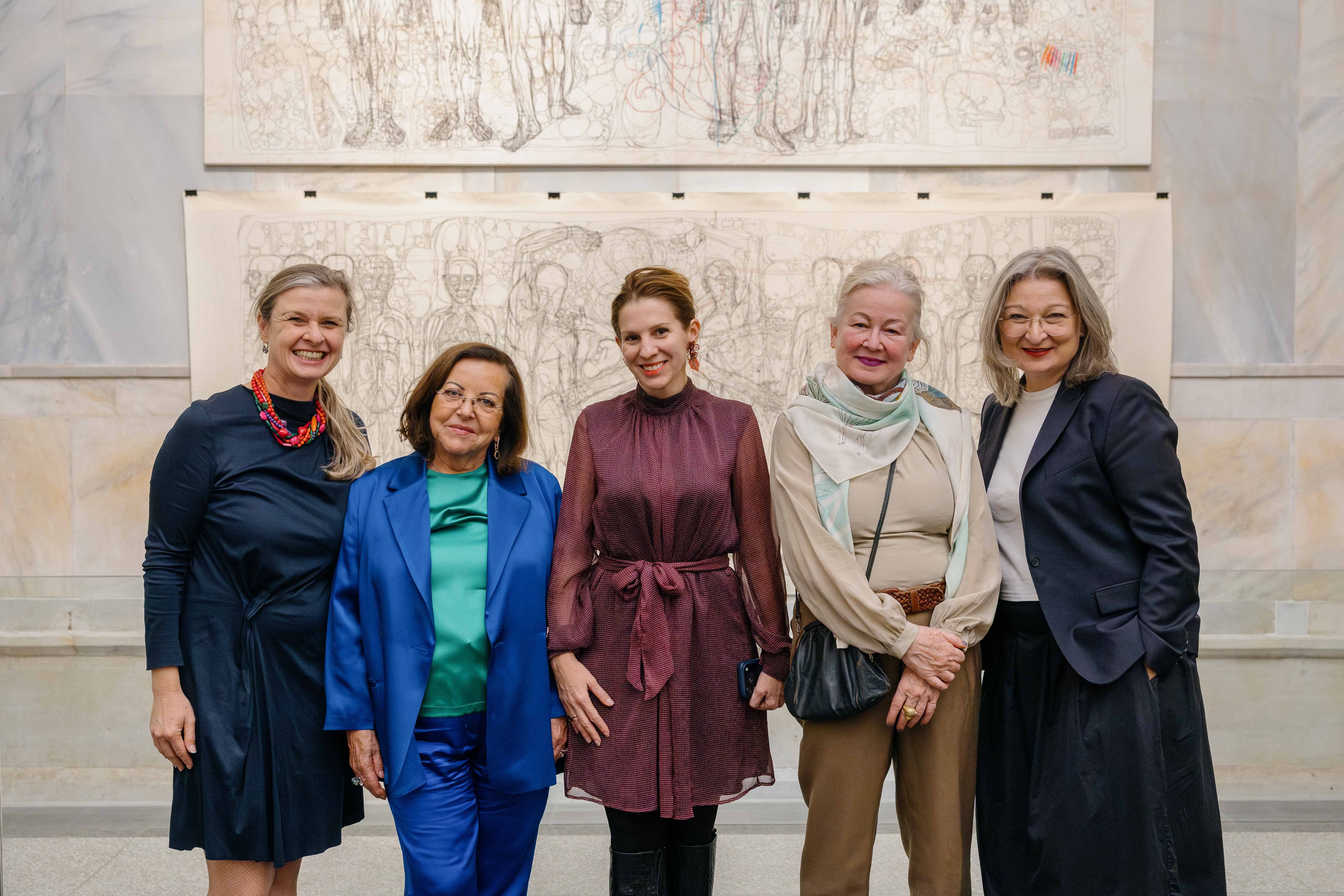 Fünf Frauen posieren lächelnd vor einer Kunstzeichnung in einem Museumsraum.