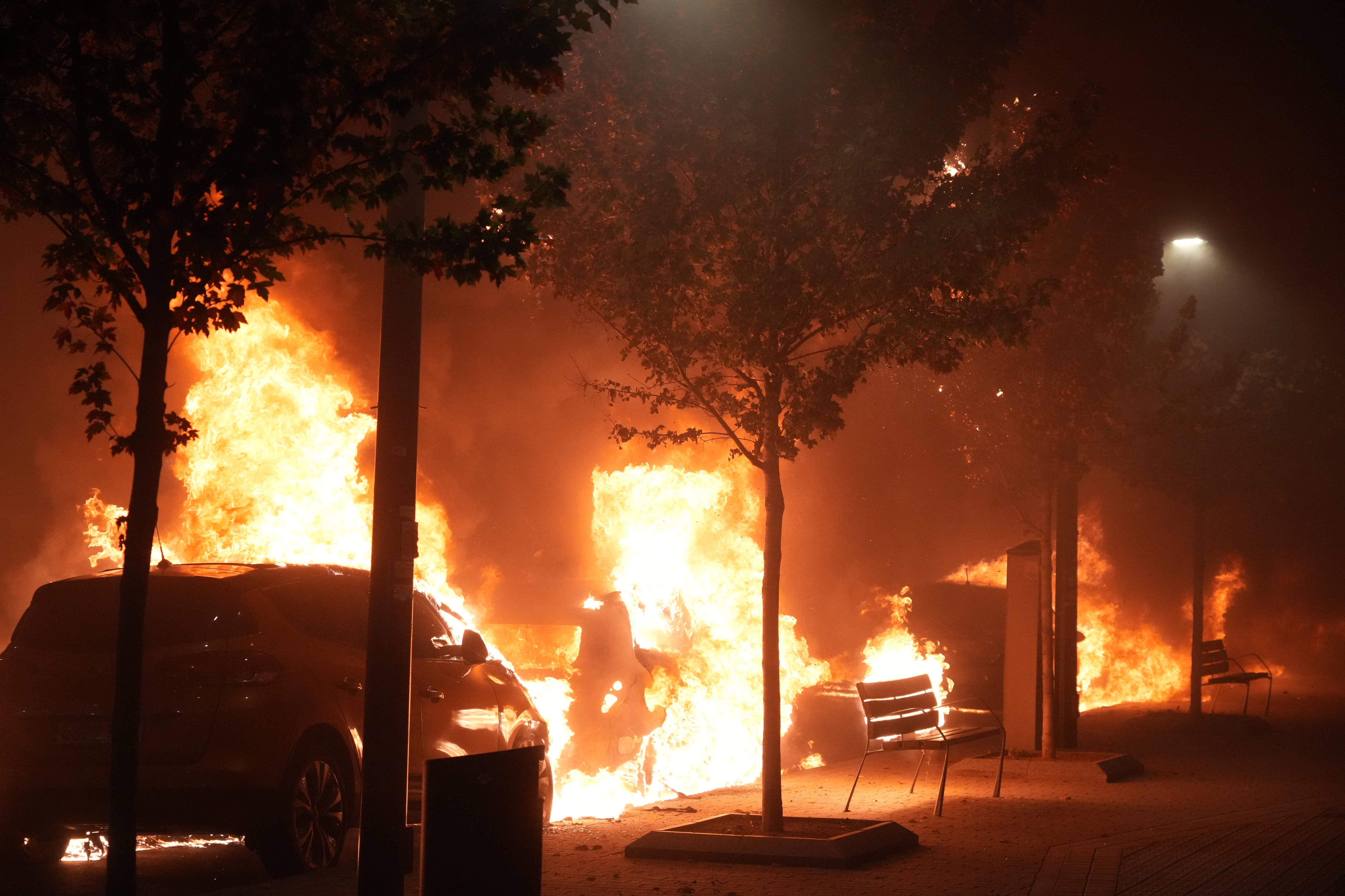 Brennende Autos und Mülleimer auf einer Straße bei Nacht, Flammen schlagen hoch zwischen Bäumen.