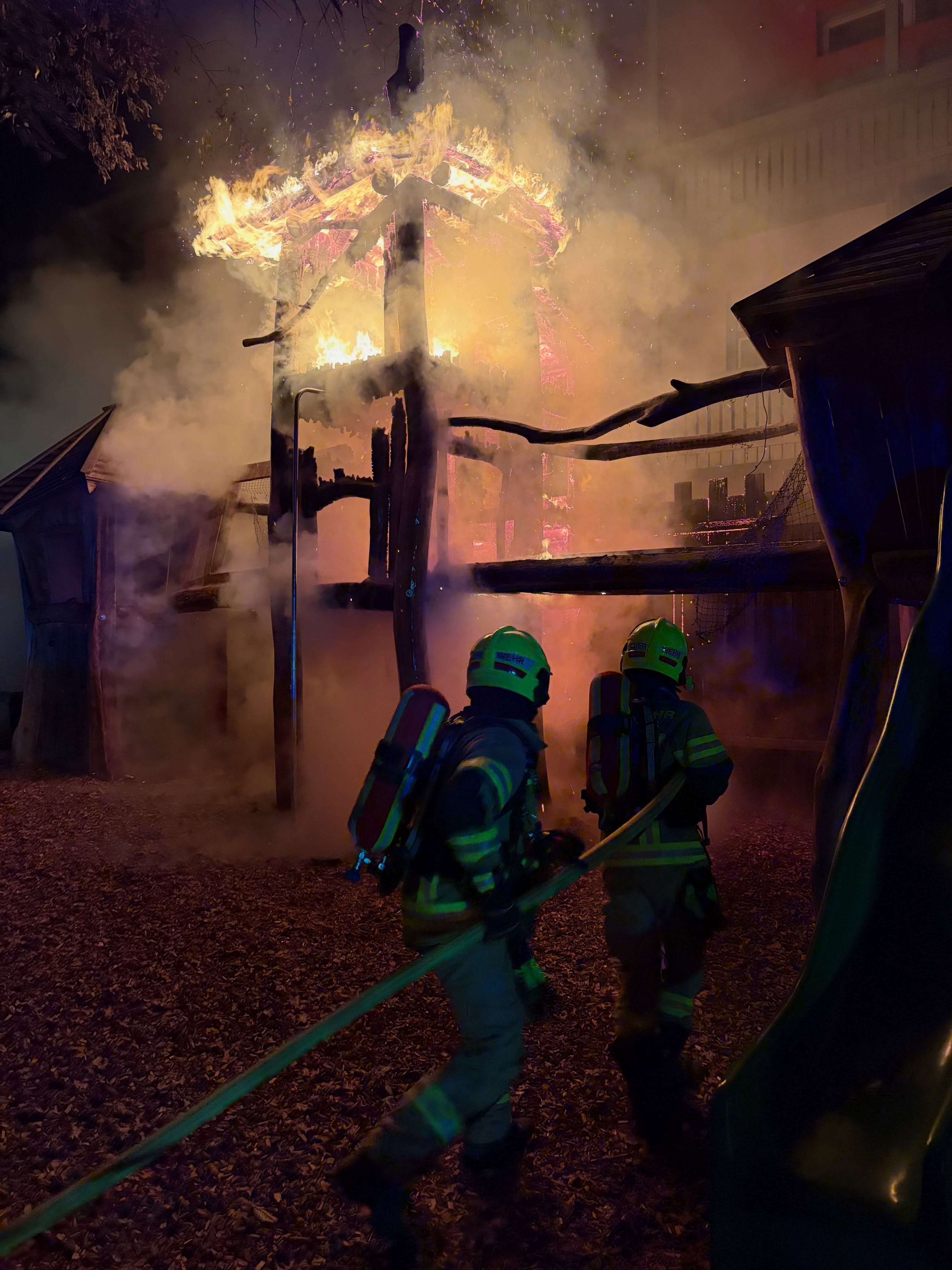 Zwei Feuerwehrleute bekämpfen einen brennenden Holzspielplatz bei Nacht mit einem Schlauch.