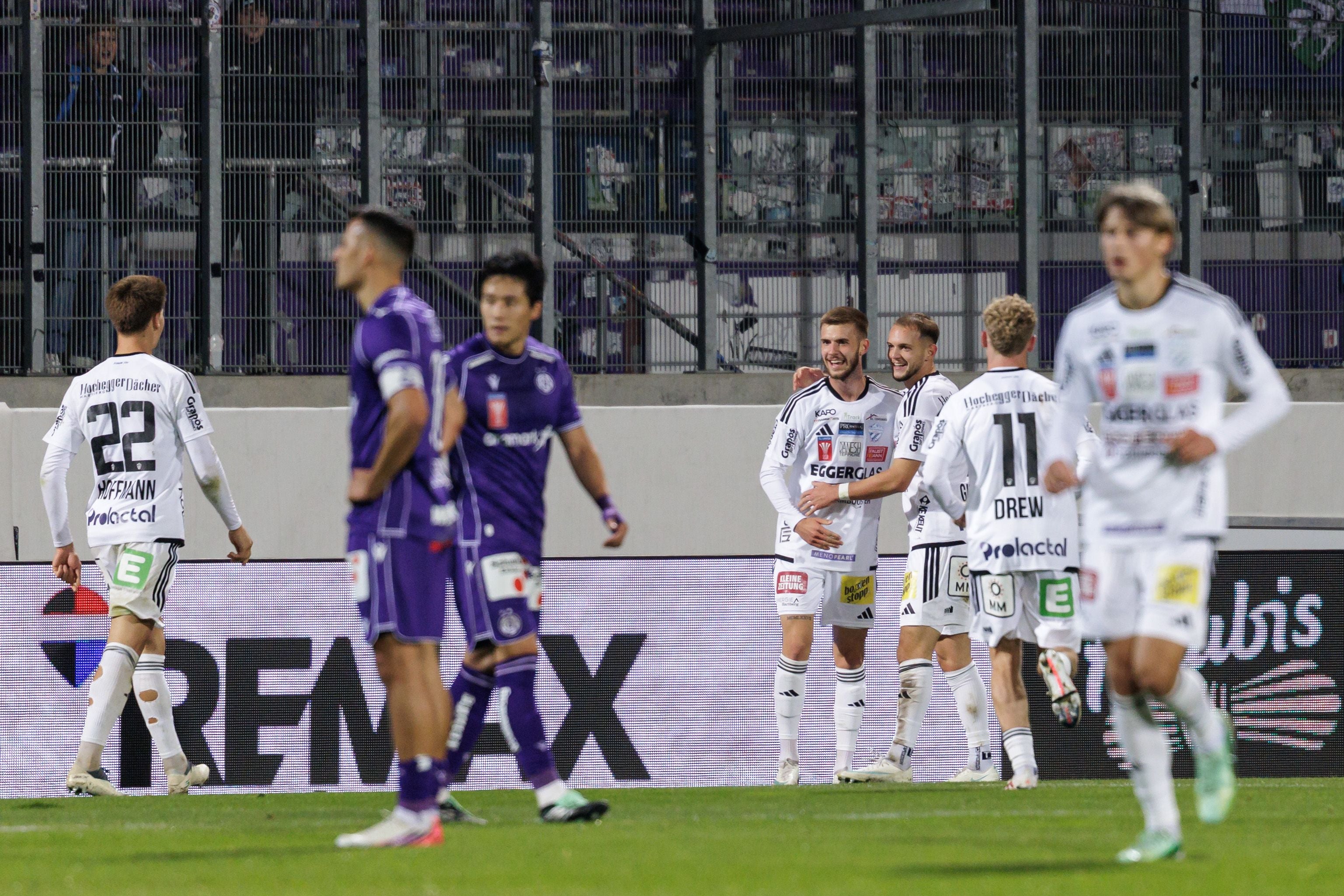Oliver Lukic feiert im weißen Trikot des TSV Hartberg gemeinsam mit seinen Teamkollegen den Treffer vor der Bandenwerbung, während die Spieler der Wiener Austria enttäuscht im lila Dress über den Platz gehen.
