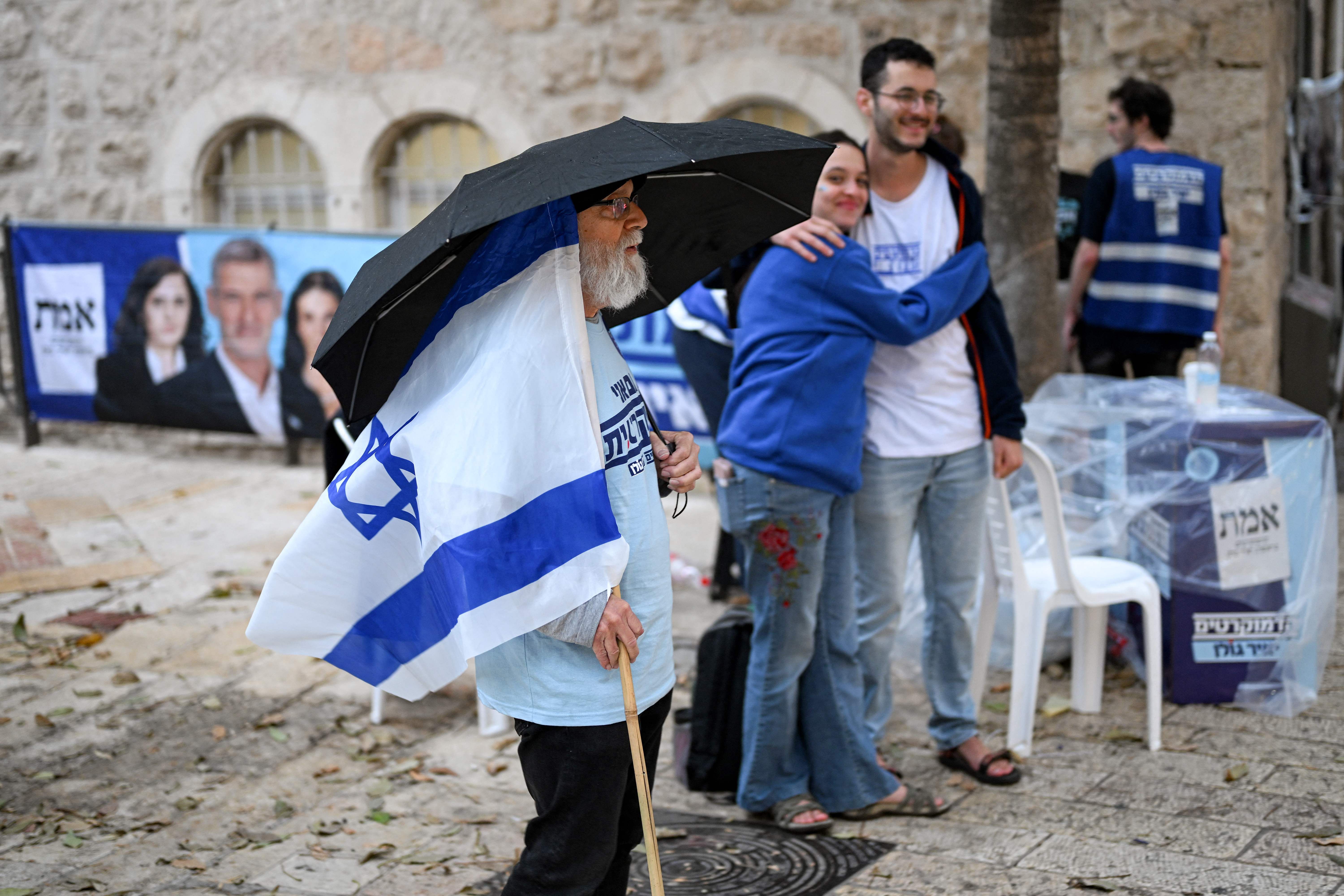 Ein Mann mit Regenschirm und israelischer Flagge steht vor einem Wahllokal in Jerusalem im Regen.